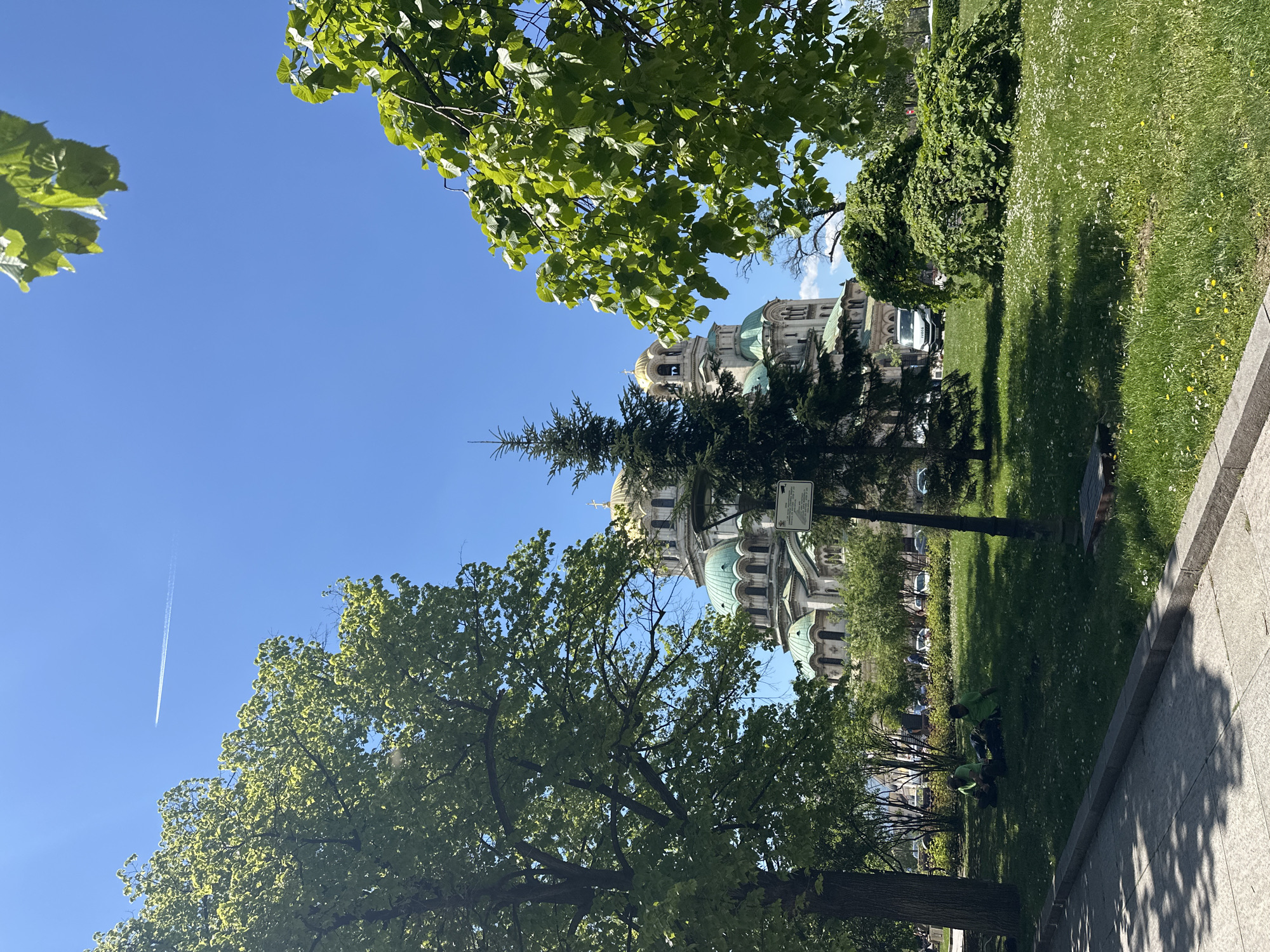 The image depicts a partial view of the Alexander Nevsky Cathedral in Sofia, Bulgaria, framed by lush green trees and a clear blue sky. A jet contrail is visible in the upper-left quadrant of the sky. The cathedral’s golden domes and green copper roof are prominent. The foreground consists of a grassy park area with some people, likely gardeners, working. The scene is captured on a sunny day.