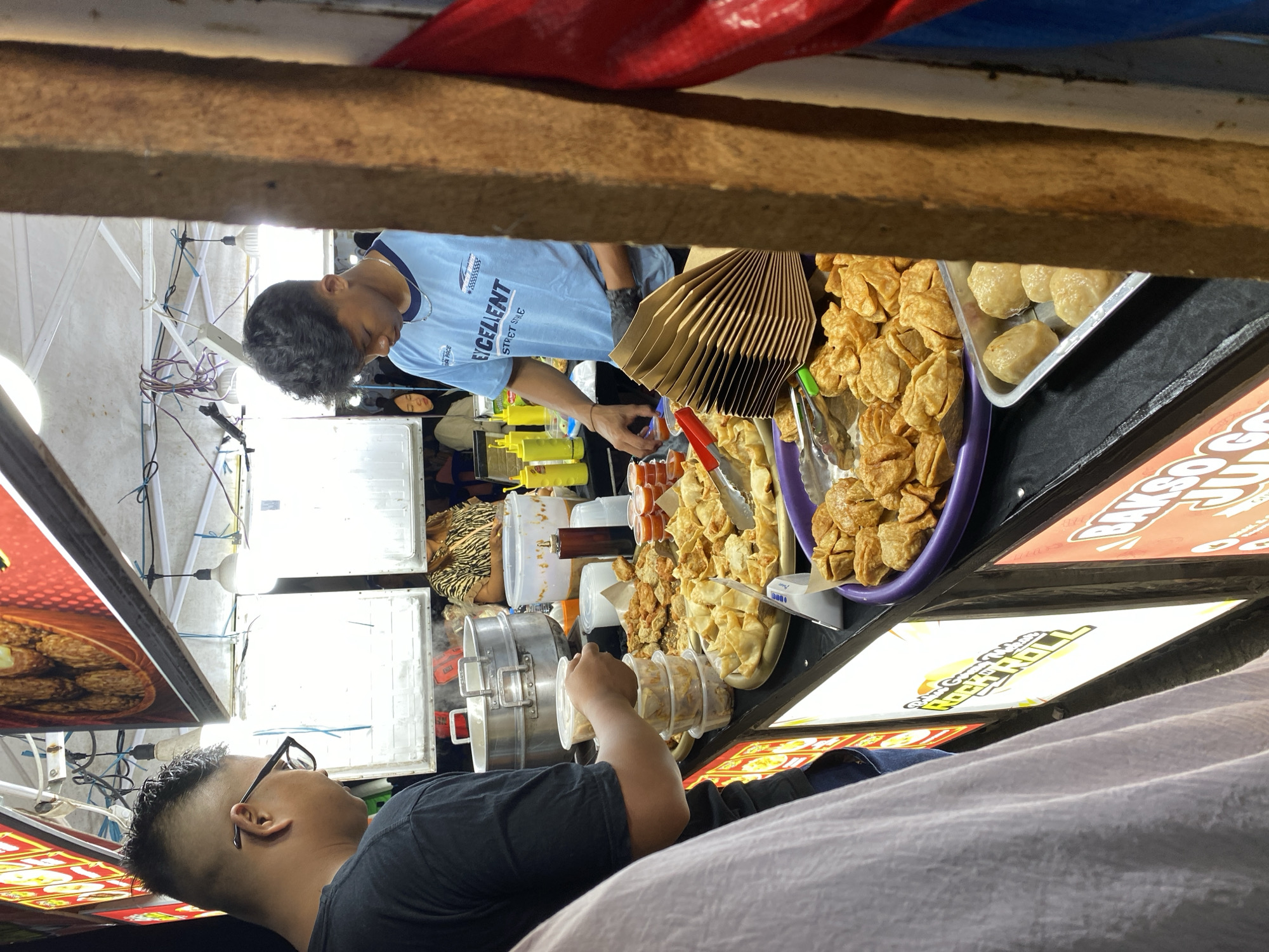 The image shows a food stall in Banjarmasin, Indonesia. The stall is selling various fried snacks, such as meatballs and dumplings. A man in a blue t-shirt is serving customers. He is wearing black gloves and holding a pair of tongs. Another man, wearing a black t-shirt and glasses, is standing in front of the stall, holding a container of food. There are several bottles of sauce and condiments on the counter, as well as a large steaming pot in the background. The stall is brightly lit, and the food is displayed attractively. The overall scene depicts a bustling street food environment.
