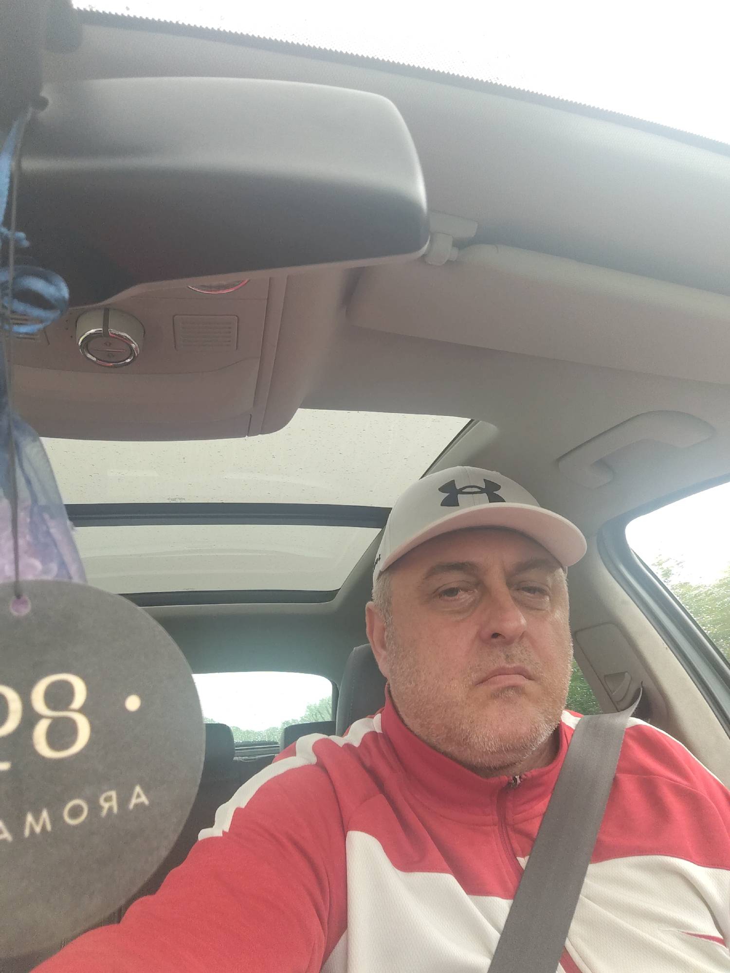 A middle-aged man with a light beard or stubble is depicted from the chest up, seated in the driver's seat of a car. He wears a light grey baseball cap featuring a dark, stylized Under Armour logo, and a red and white zippered track jacket. A grey seatbelt is fastened across his chest. His expression is neutral, appearing somewhat serious or pensive.
The setting is inside a vehicle, with a light-colored interior headliner. An overhead console is visible, equipped with a rotary dial and a small grille. Above the man, a panoramic sunroof shows a cloudy sky dotted with visible raindrops, indicating current or recent rainfall. A rearview mirror is partially visible at the top of the frame. Hanging from the rearview mirror is a circular, dark grey air freshener with the number "28" and the word "AMORA" (reversed) printed on it. Through the car windows, particularly the rear, some green foliage is discernible. The overall impression is a daytime scene inside a car during inclement weather, located in Huntingdonshire, United Kingdom.