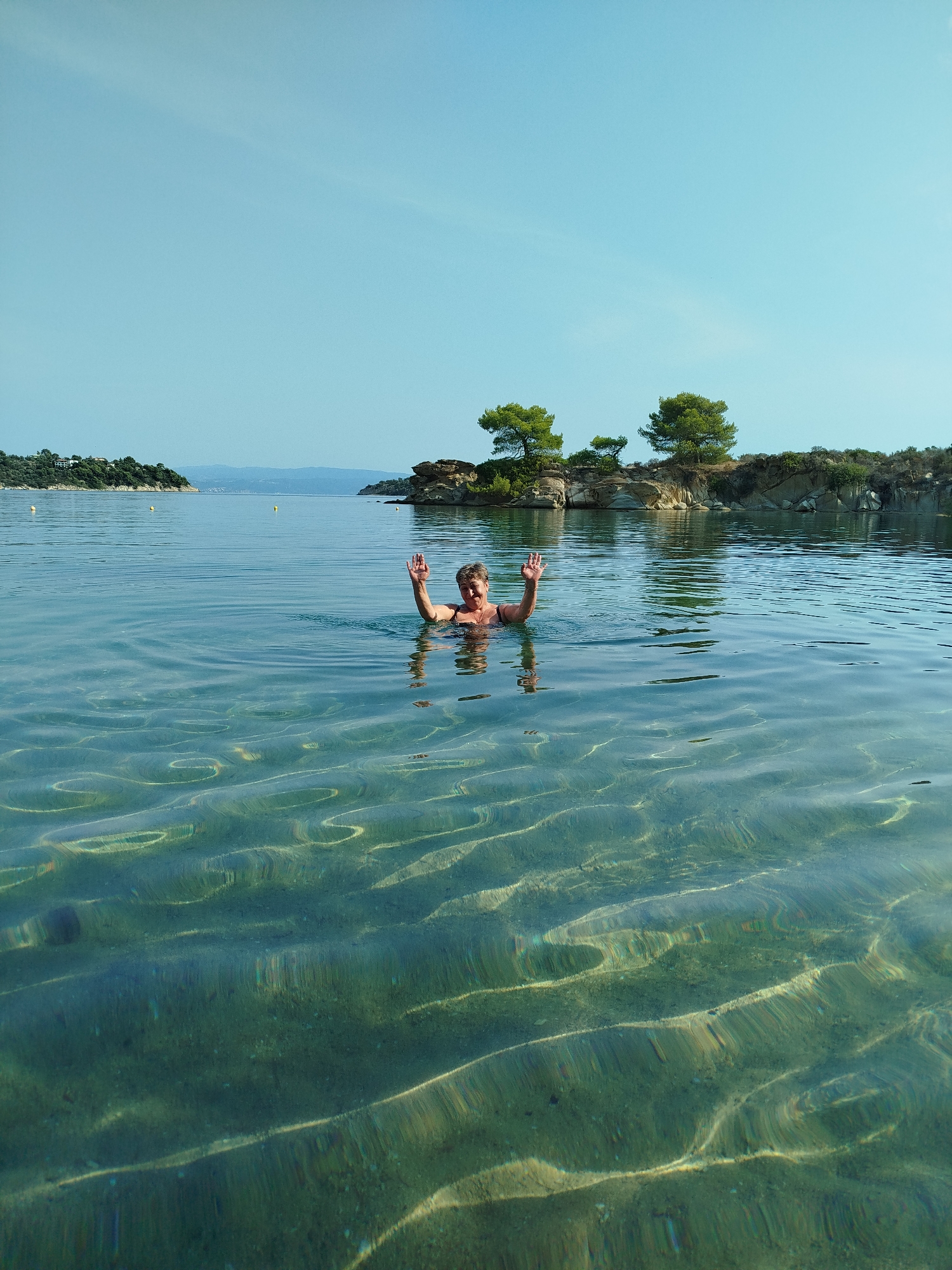A person is swimming in the clear, shallow waters of a cove in Sithonia, Greece. The person is smiling and waving with both hands, their upper body visible above the water. The seabed is visible through the translucent water, displaying rippled patterns caused by the light. In the background, a rocky shoreline with scattered pine trees and a distant landmass under a clear blue sky are visible. The water is calm and the scene suggests a serene, natural environment.