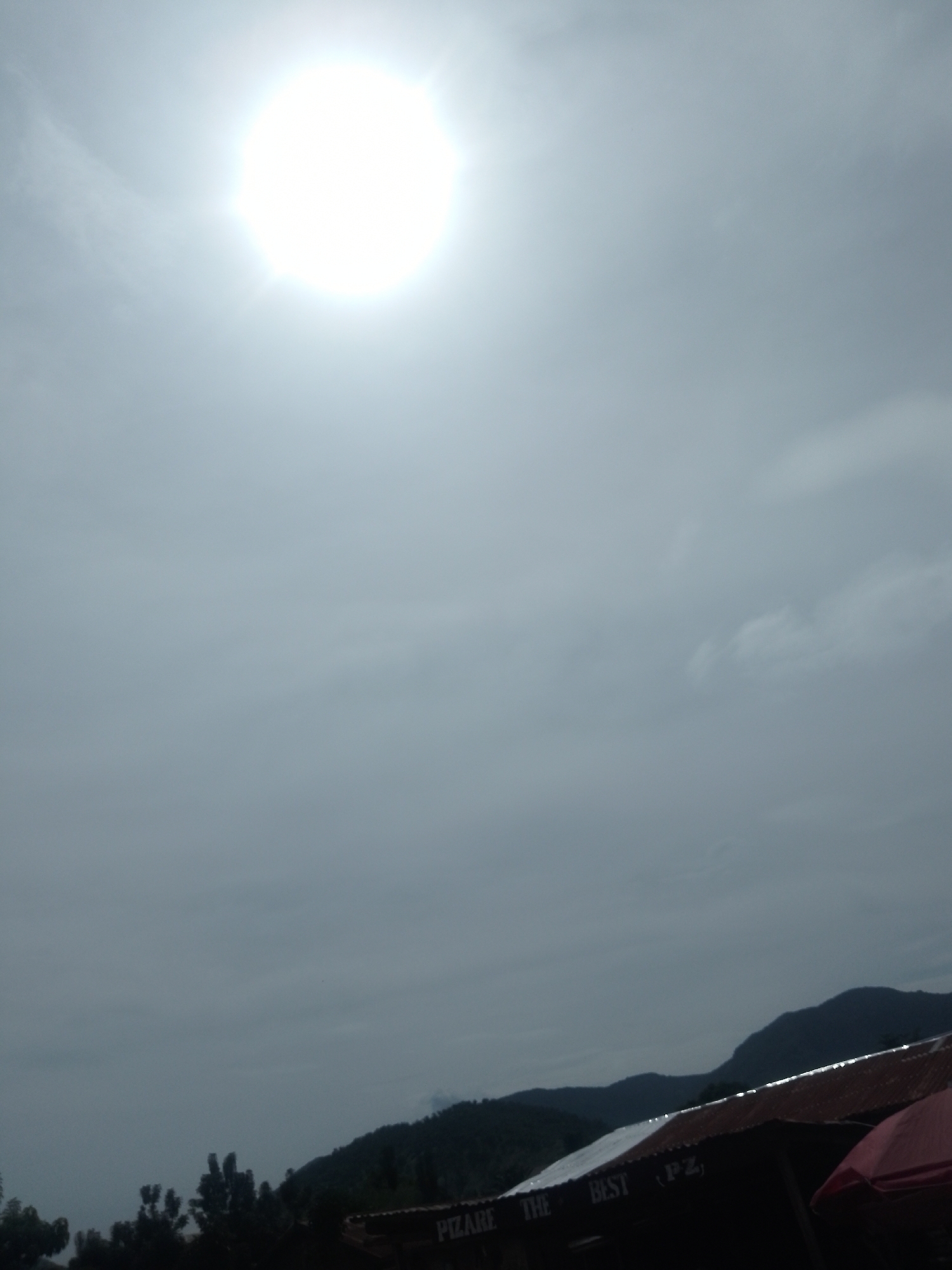 A bright, hazy sun dominates the upper portion of the image. Below, a cloudy sky transitions to a dark silhouette of trees and mountains. In the foreground, the corrugated metal roof of a building is visible, with signage that appears to read "PIZARE THE BEST (PZ)". The overall impression is one of a slightly overcast day in a somewhat rural or mountainous area, potentially a business or gathering place given the signage. The location is identified as Serti, Nigeria.