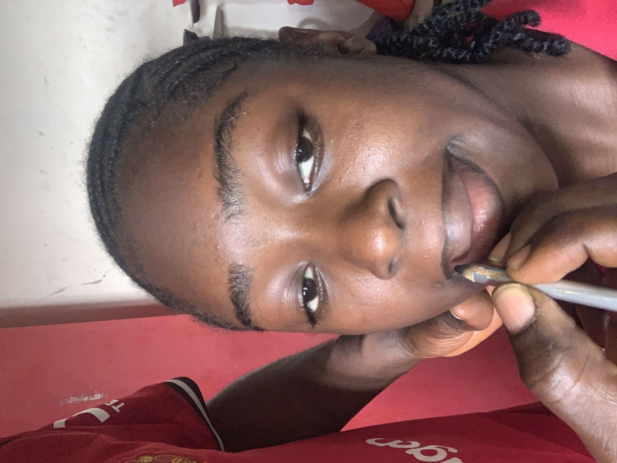 A young person, appearing to be female, is depicted in a close-up portrait, with the image rotated 90 degrees clockwise from an upright position. The setting is indoors, within the New Stadium in Nigeria. She has a dark complexion and her hair is styled in dark, neat braids (cornrows). She gazes directly towards the camera with a gentle, slightly smiling expression.
With her right hand, she holds a metallic-colored pen or pencil, bringing it to her mouth, possibly chewing or resting it against her lips. Her skin exhibits a natural sheen under what appears to be indoor lighting. She is dressed in a red t-shirt featuring white text, where the letters "TE" are clearly visible. A gold and black circular crest, highly resembling the Manchester United logo, is partially visible on the lower right side of the shirt. The background consists mainly of a red wall or surface, with some lighter, textured areas visible in the upper and left parts of the frame.