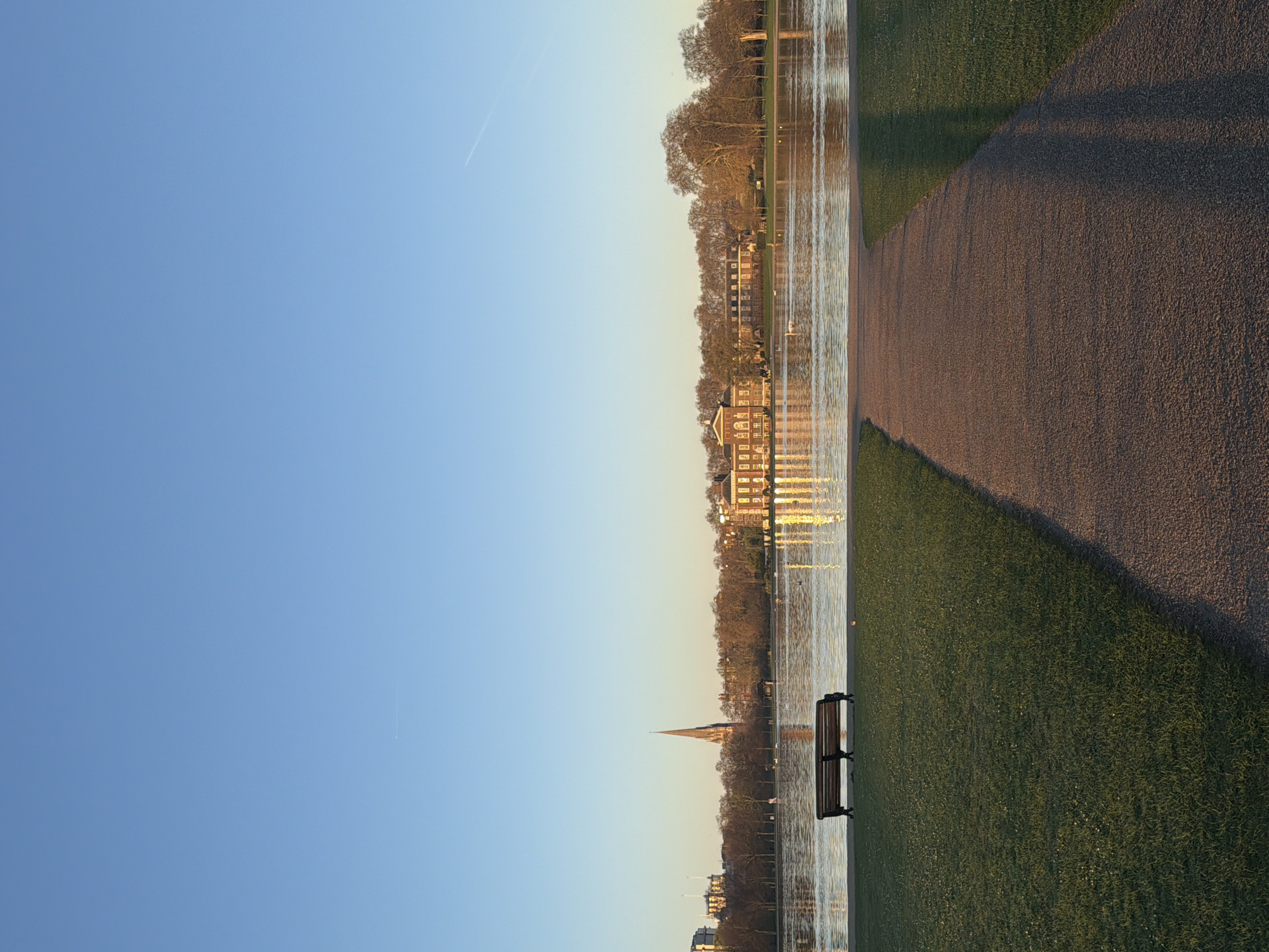 The image depicts a landscape within a London park. A pathway runs along the right side of the frame, bordered by green grass on one side and a body of water on the other. Buildings and trees are reflected in the water. A park bench sits near the water's edge. The sky is clear and blue. A contrail is visible in the sky. The location appears to be St. James's Park, London.