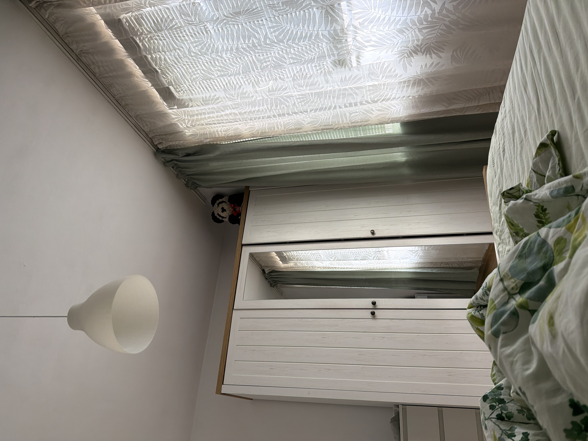 The image depicts a bedroom interior, likely in Plovdiv, Bulgaria, viewed from the perspective of lying in bed. In the foreground, a bed is covered with a duvet featuring a green and white botanical print, partially obscuring the light green bedsheets. Dominating the middle ground is a tall, white wooden wardrobe with three doors and wooden trim along the top. The middle door is a mirror, reflecting a portion of the room and the window. On top of the wardrobe, a plush toy, possibly a panda or bear, with a red bow is visible. To the right of the wardrobe are two sets of curtains covering a window. The inner curtains are a pale, sheer fabric with a white leaf pattern, while the outer curtains are a muted green. A white pendant light fixture hangs from the ceiling in the upper left quadrant of the image. In the background, a white chest of drawers with multiple drawers is partially visible to the left of the wardrobe. The lighting suggests it is daytime, with natural light filtering through the window. The room appears tidy and comfortably furnished.