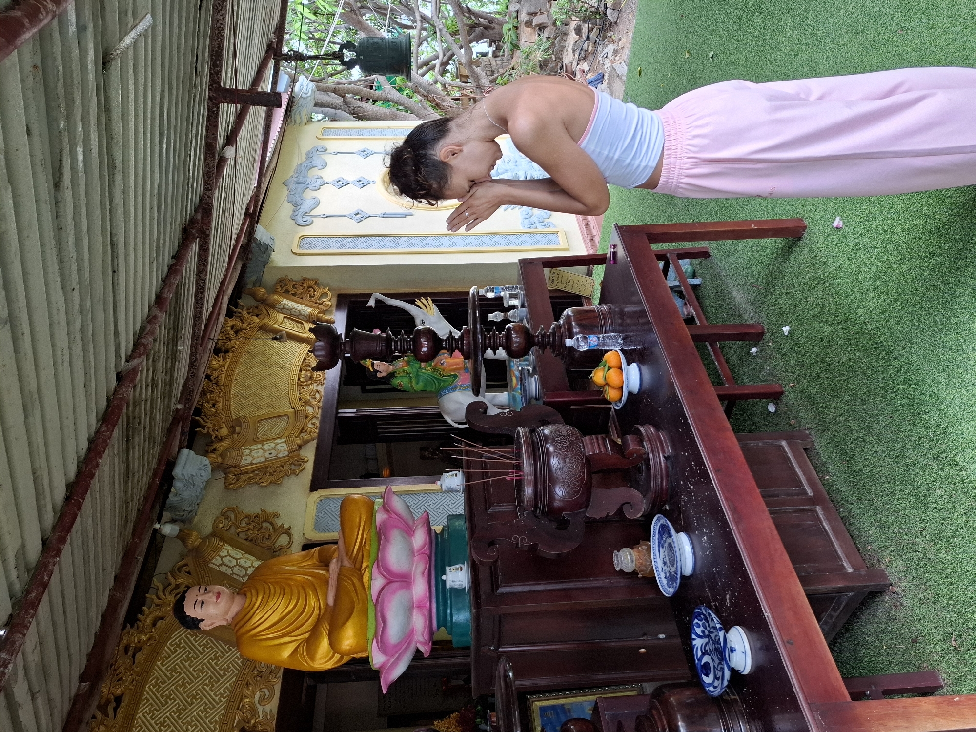 In Nha Trang, Vietnam, a young woman with dark hair styled in a bun stands with her eyes closed and hands pressed together in a gesture of prayer or meditation in front of a Buddhist shrine. She wears a white crop top and loose pink pants. The shrine features a large, golden Buddha statue seated on a lotus flower, surrounded by decorative elements. In front of the shrine, a wooden table holds an incense burner with burning incense sticks, small ceramic dishes, and a bowl of oranges. The shrine appears to be outdoors, with a corrugated metal roof overhead and lush green artificial turf covering the ground. A large bell hangs from a tree in the background.