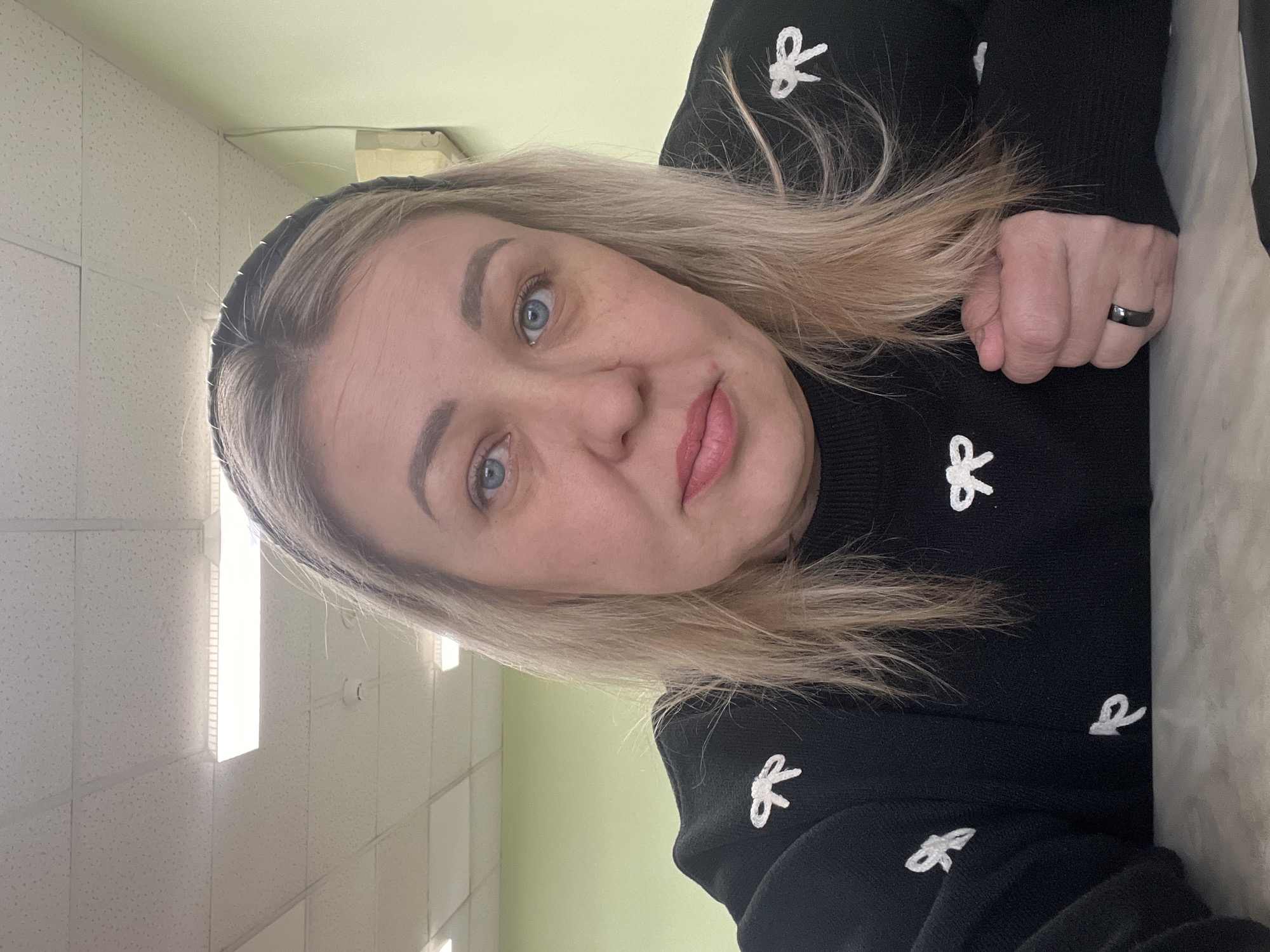 This image is a portrait of a woman, likely a selfie, taken indoors in Chaikovsky, Russia. The woman has blonde hair, styled with a black headband, and striking blue eyes. She is wearing a black long-sleeved sweater decorated with small, white bow patterns. Her expression is calm and direct, looking towards the camera, with her lips slightly parted and wearing light pink lipstick.
Her right hand is visible, clenched into a loose fist, resting on what appears to be a light-colored surface or fabric next to her. She is wearing a simple black ring on the ring finger of her right hand.
The setting appears to be an indoor environment, possibly an office, classroom, or public building. The background features a white ceiling with square acoustic tiles and bright, rectangular fluorescent light fixtures, suggesting ample artificial illumination. A light green wall is also visible in the background. The overall lighting is bright and even, typical of an indoor daytime setting. There is no discernible text in the image. The photo is rotated 90 degrees clockwise.
