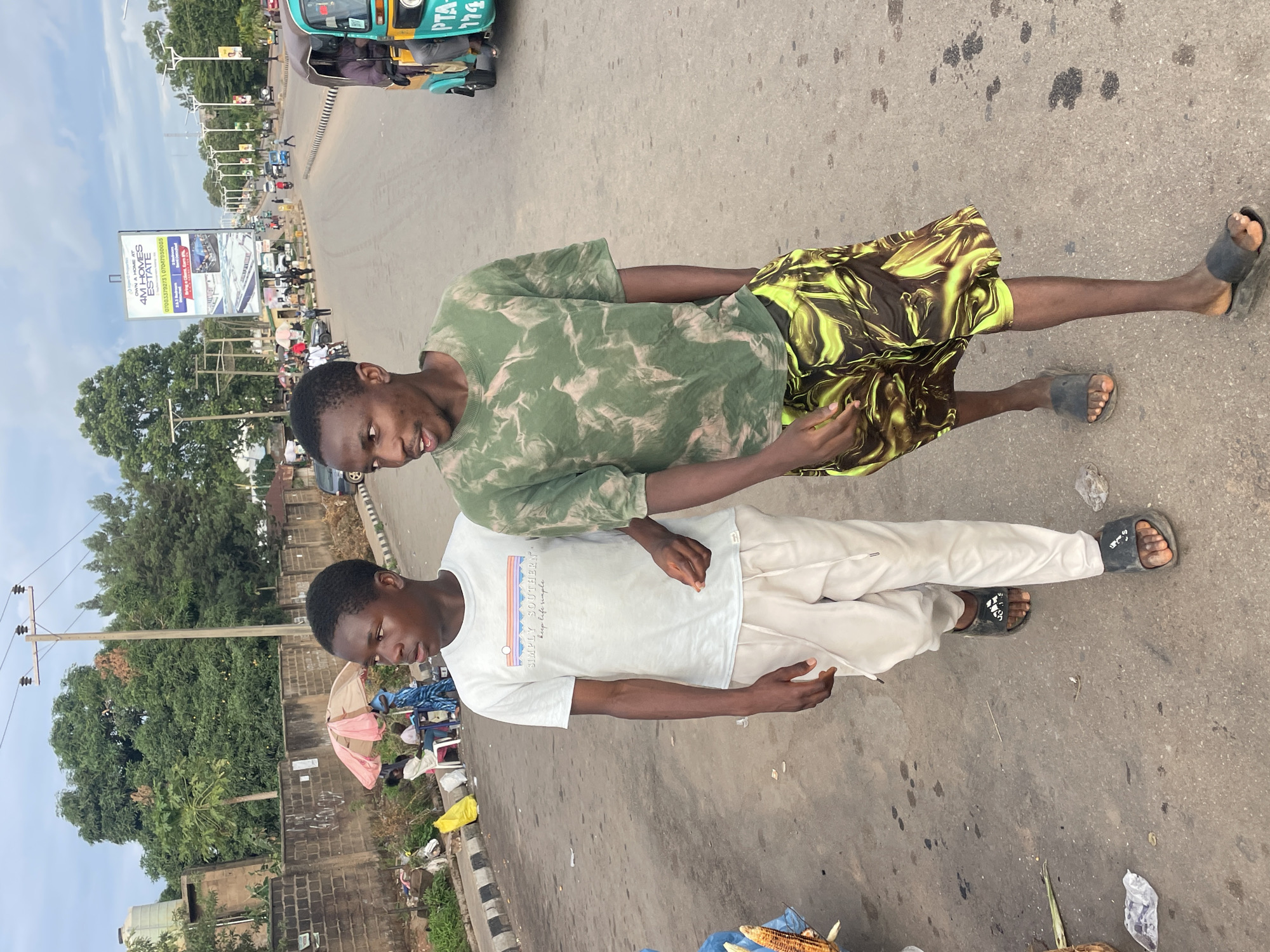 This image depicts a street scene in Jos, Plateau State, Nigeria. Two young Black males walk leftward on an asphalt road. The male on the left wears an off-white short-sleeved t-shirt with "SIMPLY SOUTHERN" text, light-colored drawstring pants, and black Adidas-branded slide sandals; he holds an unlit cigarette in his right hand and looks to his left. The male on the right, slightly ahead, wears a green and light-patterned short-sleeved t-shirt, dark shorts with a vibrant yellow/green abstract pattern, and dark slide sandals; he faces left with his mouth open, indicating speech or reaction.
On the left side of the road, a street vendor operates under a pink and beige umbrella, with individuals seated on white plastic chairs nearby. Yellow plastic bags are visible on the ground beside a concrete block wall. A white water tank sits atop a structure behind this wall. In the upper-right background, a large billboard for "4M HOMES ESTATE" by "Super G Properties" is visible, displaying contact numbers 0703379273 and 07047950003. Further down the road, a blue and yellow motorized tricycle taxi, commonly known as a Keke Napep, with a partial license plate "PTA 174" is visible, moving away. The road is wide, appearing to have multiple lanes or a central median with streetlights in the distance. Various other vehicles and pedestrians are present further along the road. The environment includes abundant green trees under an overcast sky. The road surface shows signs of wear, and some litter is present on the ground.