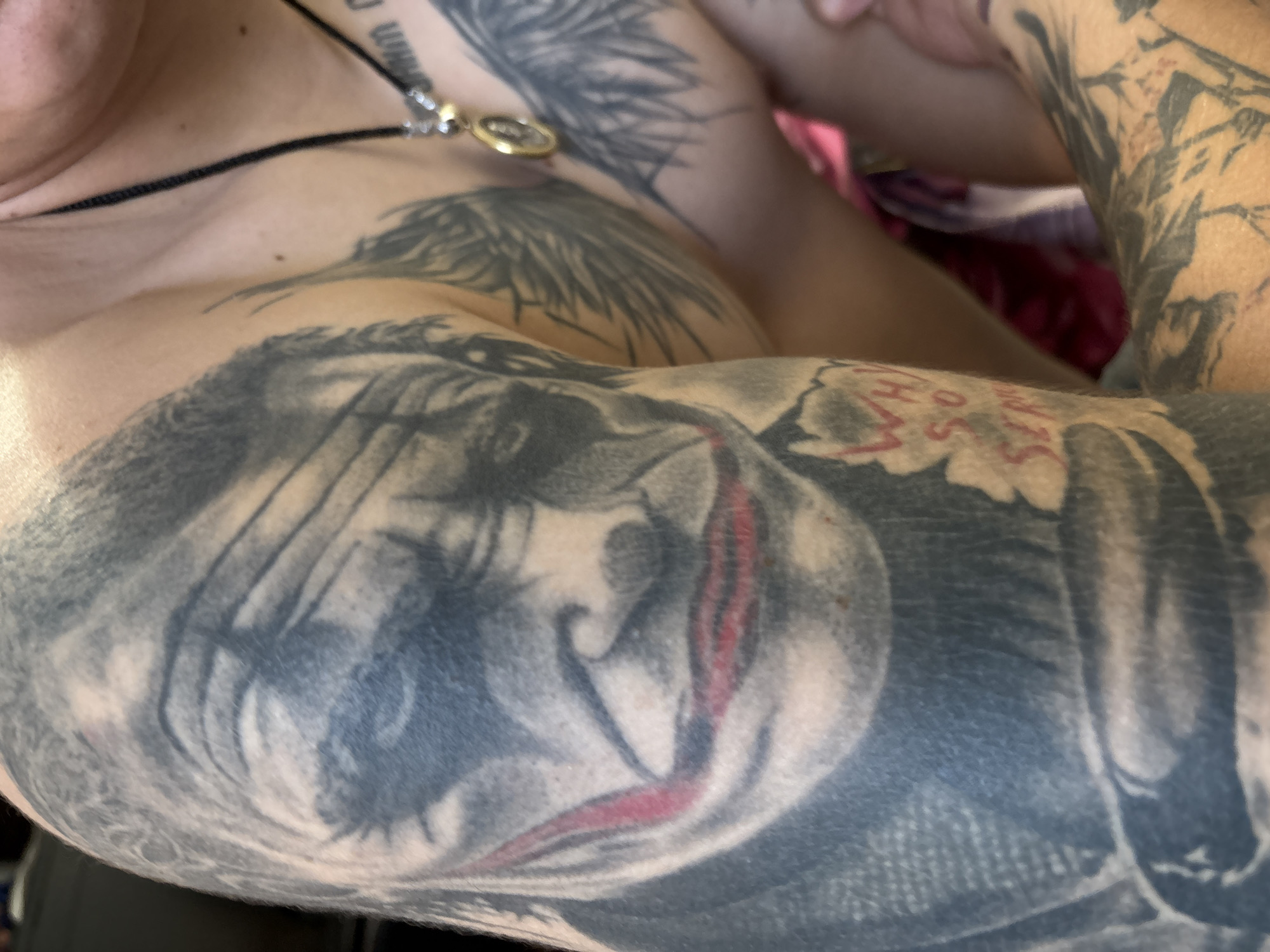The image displays a close-up of a person's left shoulder and upper arm, heavily tattooed. The most prominent tattoo is a detailed portrait of the Joker from "The Dark Knight," featuring his signature makeup and a portion of his red smile. To the right of the Joker tattoo, on the person's chest and upper arm, are tattoos of what appear to be feathered wings and a circular pendant. Further down the arm, there are visible tattoos of stylized lettering and floral elements. The skin is fair, and some areas show slight redness, possibly indicating recent tattooing or irritation. The background is blurred but suggests an indoor setting with ambient lighting. The image focuses on the tattoos, with the person's face and body mostly obscured.