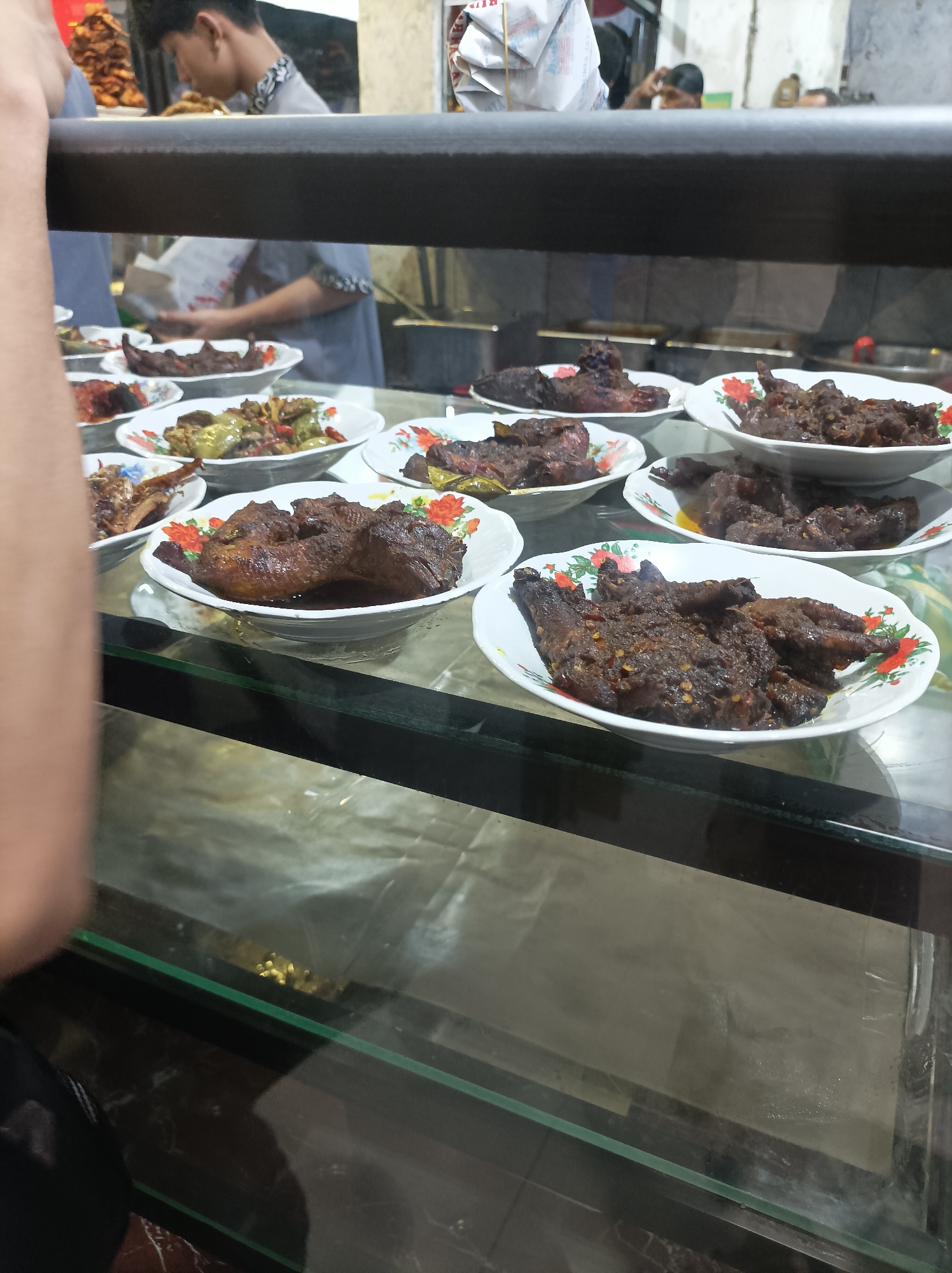 A close-up, eye-level shot shows a display case filled with various Indonesian dishes, likely at a warung makan in West Jakarta, Indonesia. The focus is on the food, presented in white ceramic bowls with floral patterns. Visible dishes include what appears to be rendang, fried chicken, and other meat dishes. In the background, blurred figures of people are visible, including a young man in a grey shirt with patterned trim. The lighting suggests an indoor setting, possibly a market or a casual eatery. The display case has a dark, reflective surface at the bottom, and a thick, dark grey horizontal bar runs across the top of the frame, obscuring some of the background.