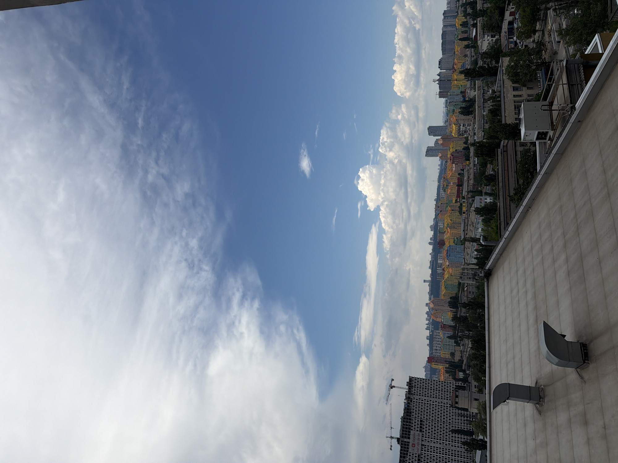 The image shows a cityscape in Kyiv, Ukraine, from a high vantage point, likely a rooftop. The foreground is dominated by a flat, grey rooftop with two large, grey ventilation pipes. Beyond the rooftop, a sprawling urban landscape unfolds under a partly cloudy blue sky. In the mid-ground, a large, unfinished building with a grid-like facade is under construction, with several cranes visible. Adjacent to it, a densely packed residential area features buildings with brightly colored facades in shades of yellow, orange, pink, and red, creating a vibrant contrast with the more muted tones of other structures. A distant skyline of taller buildings marks the city's center. Trees are scattered throughout the city, providing pockets of green. The overall scene depicts a mix of ongoing development and established urban life.