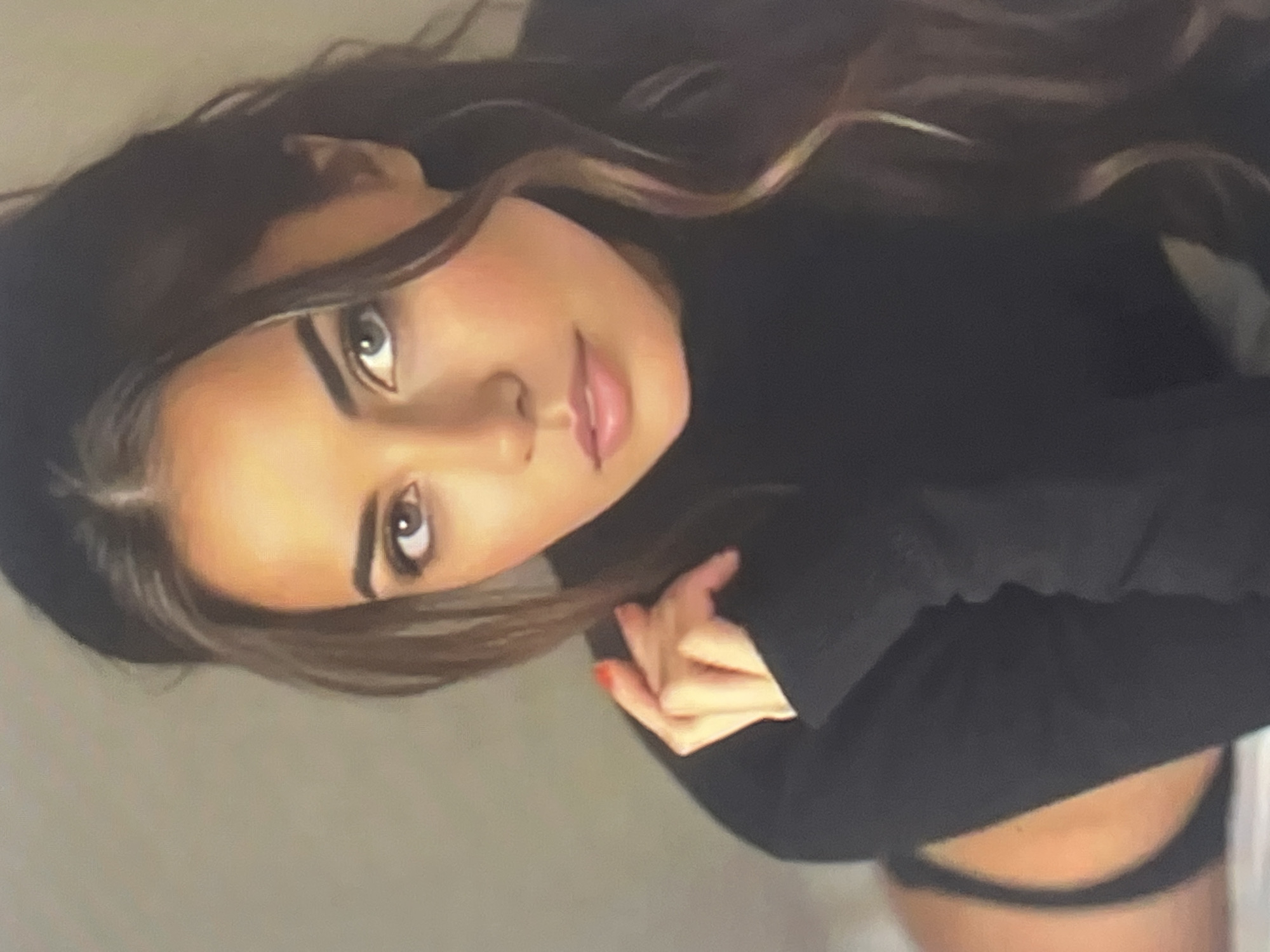The image shows a close-up of a young woman with long, dark, wavy hair, a dark top, and what appears to be black underwear or bikini bottoms visible at the bottom left of the frame. She has fair skin, dark eyebrows, and her eyes are looking towards the viewer. Her left hand is raised to her face, with red nail polish visible on one finger. The background is a plain, light-colored wall. There is no discernible location in the city of Qarağandı city administration, Kazakhstan in this image.