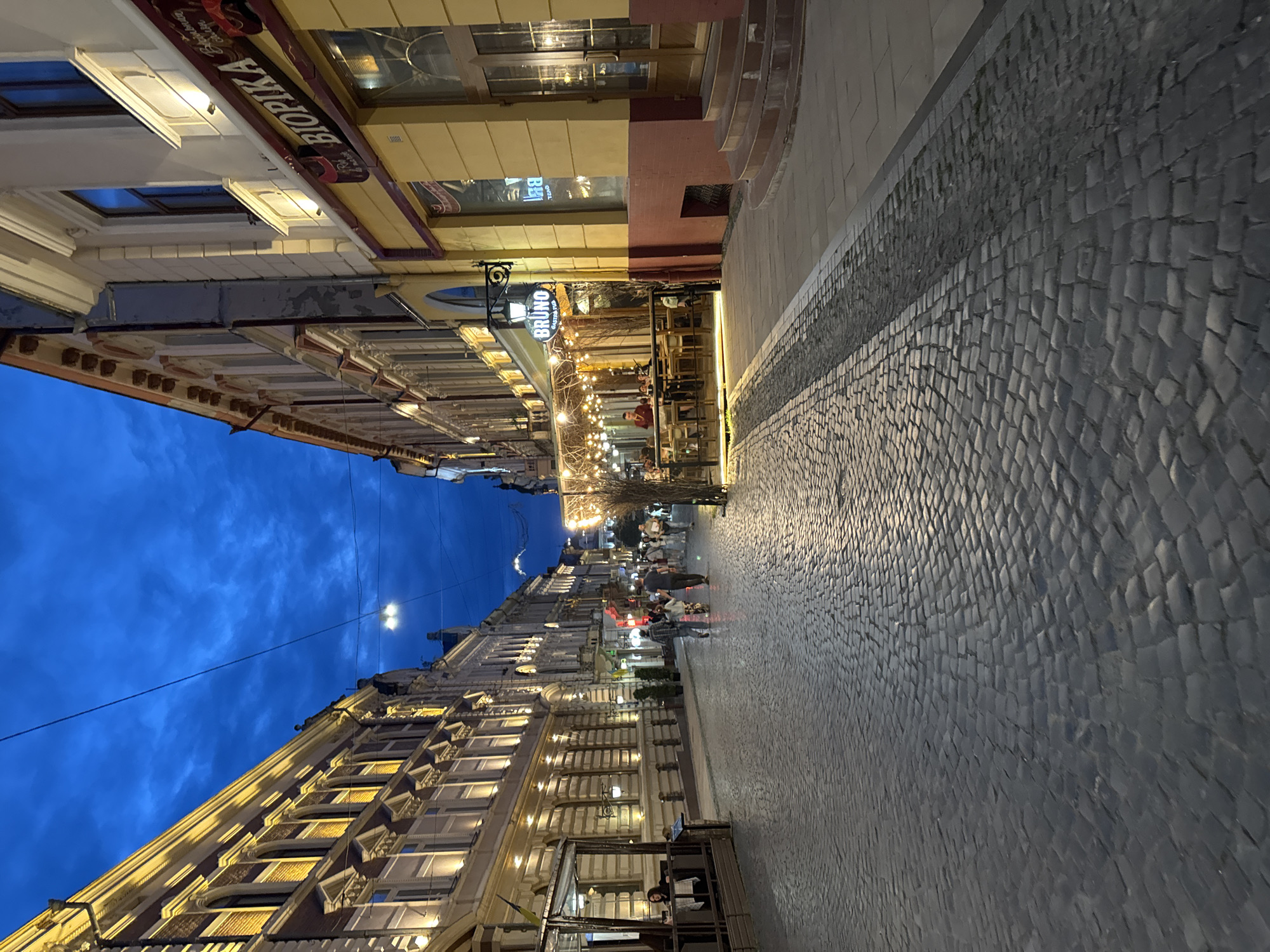 The image depicts a cobblestone street in Chernivtsi, Ukraine, at dusk or early evening. The sky is a deep blue, with some wispy clouds visible. The street is lined on both sides by historic buildings with ornate facades, illuminated by warm, artificial lights. Many windows in the buildings are lit from within.
On the left side of the street, a building with a yellow and white facade features arched windows and decorative moldings. An outdoor seating area for a cafe or restaurant, enclosed by glass, is visible on the ground floor. A woman is seated at a table inside this area.
On the right side of the street, a yellow building with a red lower section has a prominent sign reading "BIOPIKA." Another building further down the street has a sign for "BRUNO BISTRO & BAR," with string lights illuminating an outdoor seating area where several patrons are visible. Further down the street, more people are walking, and lights from other establishments are visible.
The cobblestone street is wet in places, reflecting the lights. People are seen walking in small groups, suggesting a leisurely evening stroll. The overall atmosphere is that of a charming European city street at twilight, with a sense of activity and ambiance from the illuminated businesses.