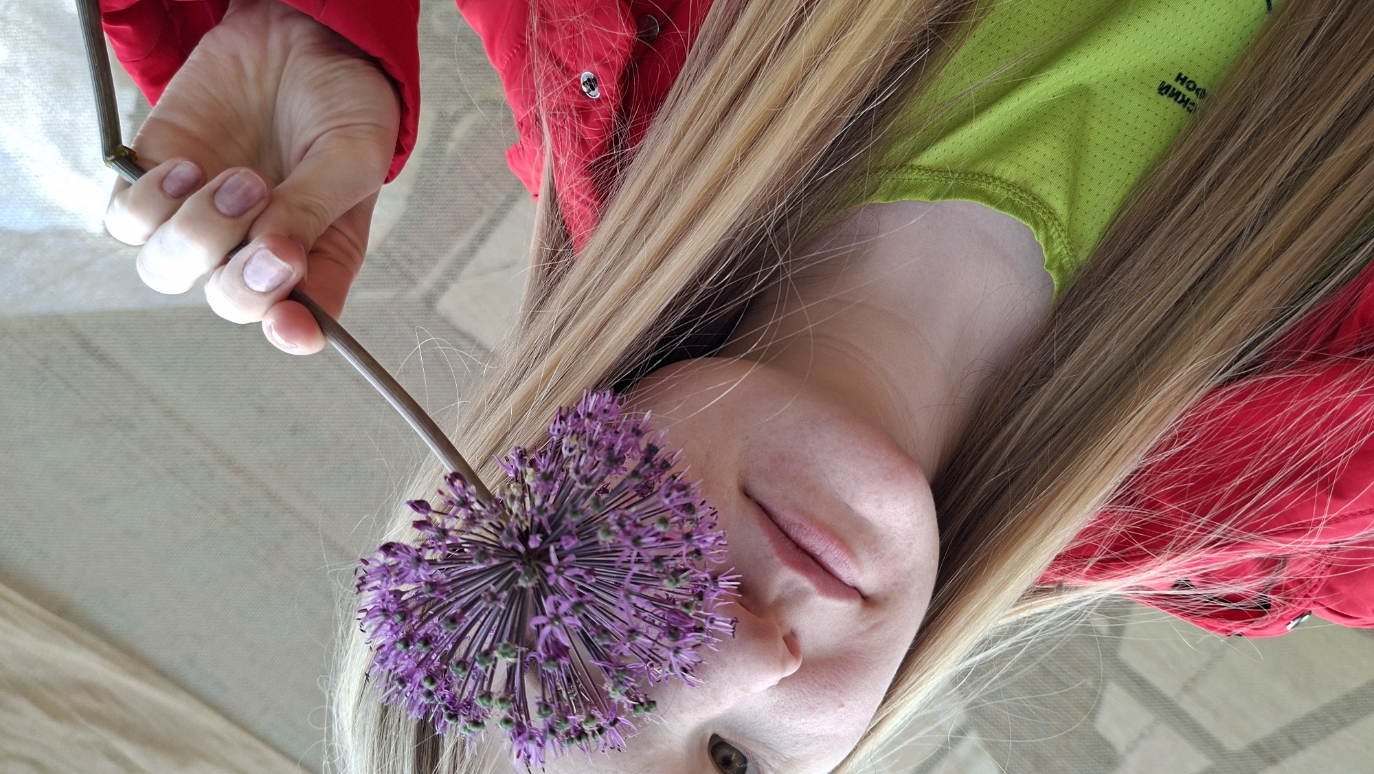 A woman is lying down, holding a purple allium flower over her face. The flower is positioned so that it covers her left eye. She has fair skin, blond hair with darker roots, and is wearing a red jacket over a green shirt. Her hand is visible holding the stem of the flower. The background appears to be a light-colored surface. There is no indication of location or city, Большой Букор, Russia, visible in the picture.