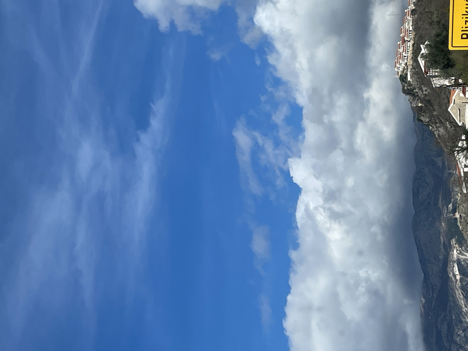 The image depicts a mountainous landscape under a dynamic sky in Blizikuće, Montenegro. The upper two-thirds of the frame are dominated by a clear, bright blue sky with scattered wispy cirrus clouds. Below this, a dense layer of white and grey cumulus clouds extends across the mid-ground, casting shadows that darken the distant mountain range on the left. On the right side, a rocky hillside rises, topped by a cluster of buildings featuring white facades and prominent reddish-orange tiled roofs. More white-walled structures are visible lower down on the slopes, with sparse, bare deciduous vegetation interspersed among the rocks. In the bottom right corner, a yellow road sign with black lettering partially reads "Blizikuće", confirming the location. No individuals, vehicles, or discernible activities are present in the scene.