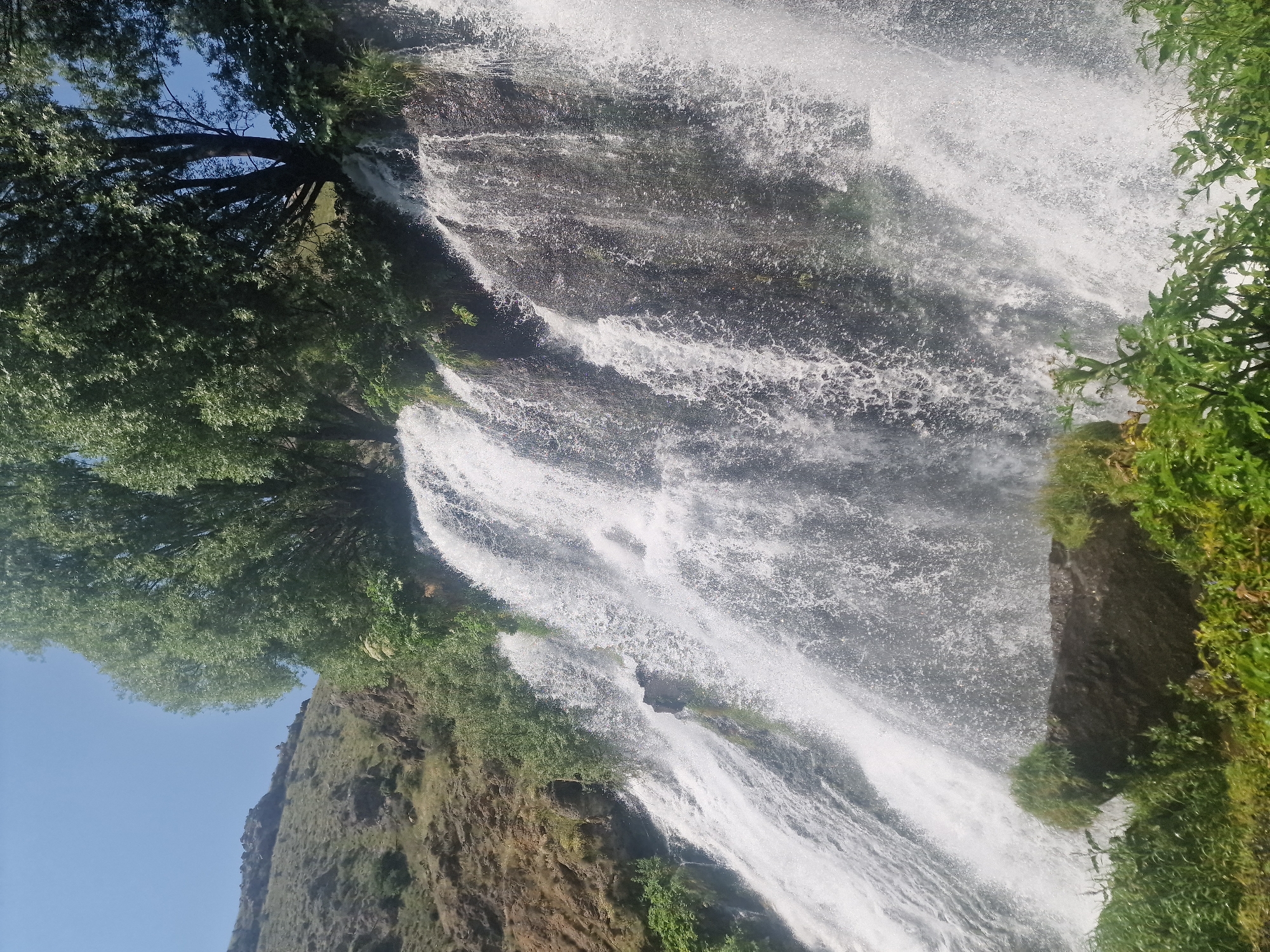 The image depicts a powerful waterfall cascading down a rocky cliff face, with lush green vegetation and trees framing the scene. The water is a torrent of white spray, creating a dynamic and energetic atmosphere. In the foreground, the rocks are covered in vibrant green moss and plants, suggesting a damp and fertile environment. The cliff itself is a mixture of dark rock and lighter-colored earth, with some patches of scrubby vegetation. Above the waterfall, large trees with dense foliage reach towards a clear blue sky. The overall impression is one of natural beauty and raw power. This is the Jermuk Waterfall, located in the city of Jermuk, Armenia, which is situated in the province of Vayots Dzor, not Sisian.