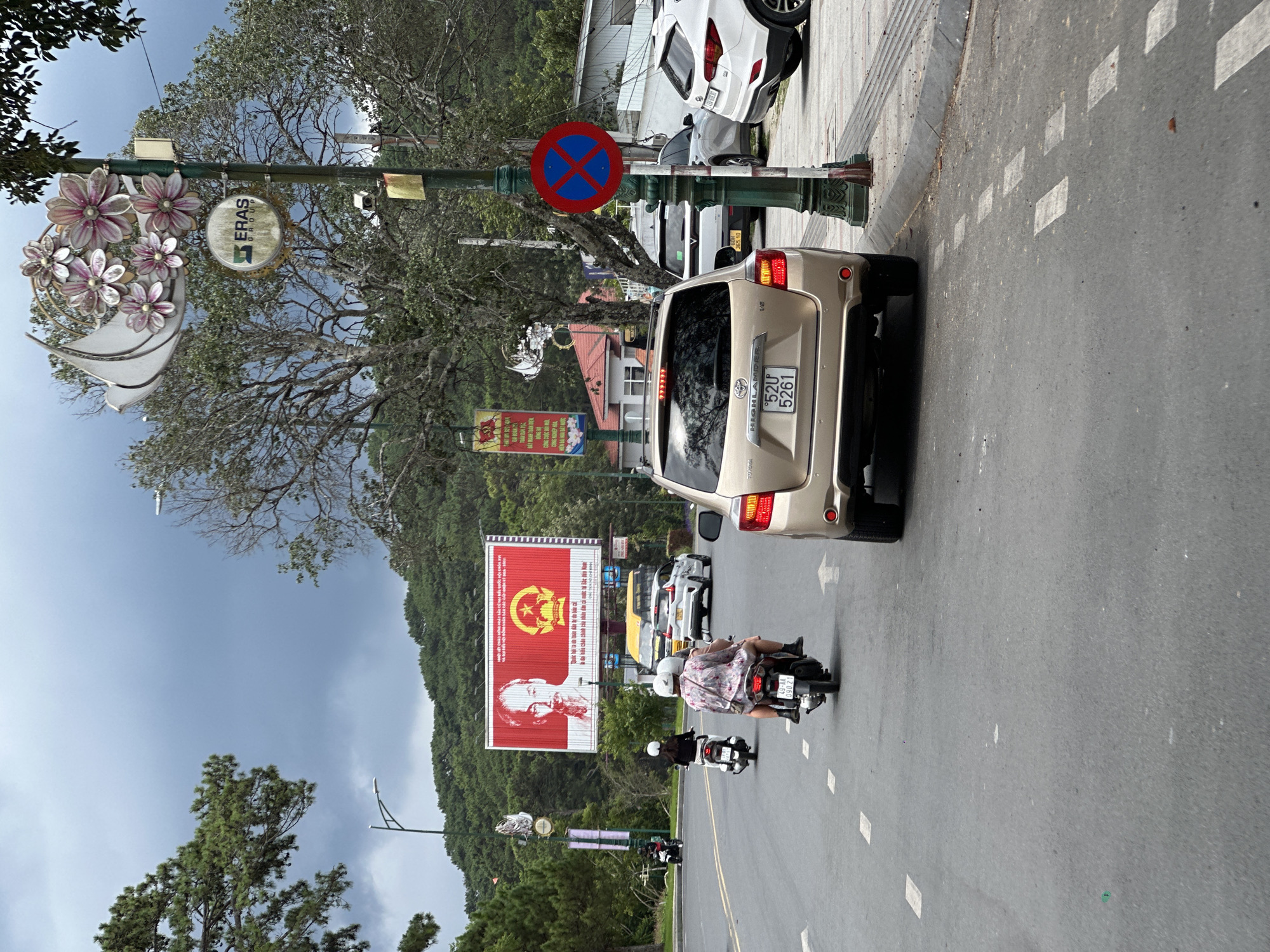 The image captures a street scene in Da Lat, Vietnam, under a cloudy sky. The foreground features the rear of a gold Toyota Highlander SUV with license plate '52U 5261', parked on the right side of the road. To the right of the SUV, a green lamppost displays a red and blue circular 'no parking' sign. The road is asphalt with white lane markings.
In the middle ground, a two-lane road is visible with traffic. Two motorbikes are in motion. The first motorbike, to the left, is ridden by a person wearing a helmet and a white shirt, and has a passenger wearing a helmet and a floral-patterned shirt. They are moving away from the camera. Further ahead, another motorbike with a rider in a white helmet is also moving away. To the right of the road, parked cars include a white SUV and a lighter-colored SUV. Beyond the road, houses with red roofs are visible amongst lush green trees and rolling hills in the background.
A large billboard is prominently displayed on the left side of the road, featuring a portrait of Ho Chi Minh and the Vietnamese national emblem. Decorative street lights with flower motifs are visible along the street. The overall environment suggests a city street with a mix of residential and commercial elements, set against a backdrop of natural greenery typical of Da Lat.