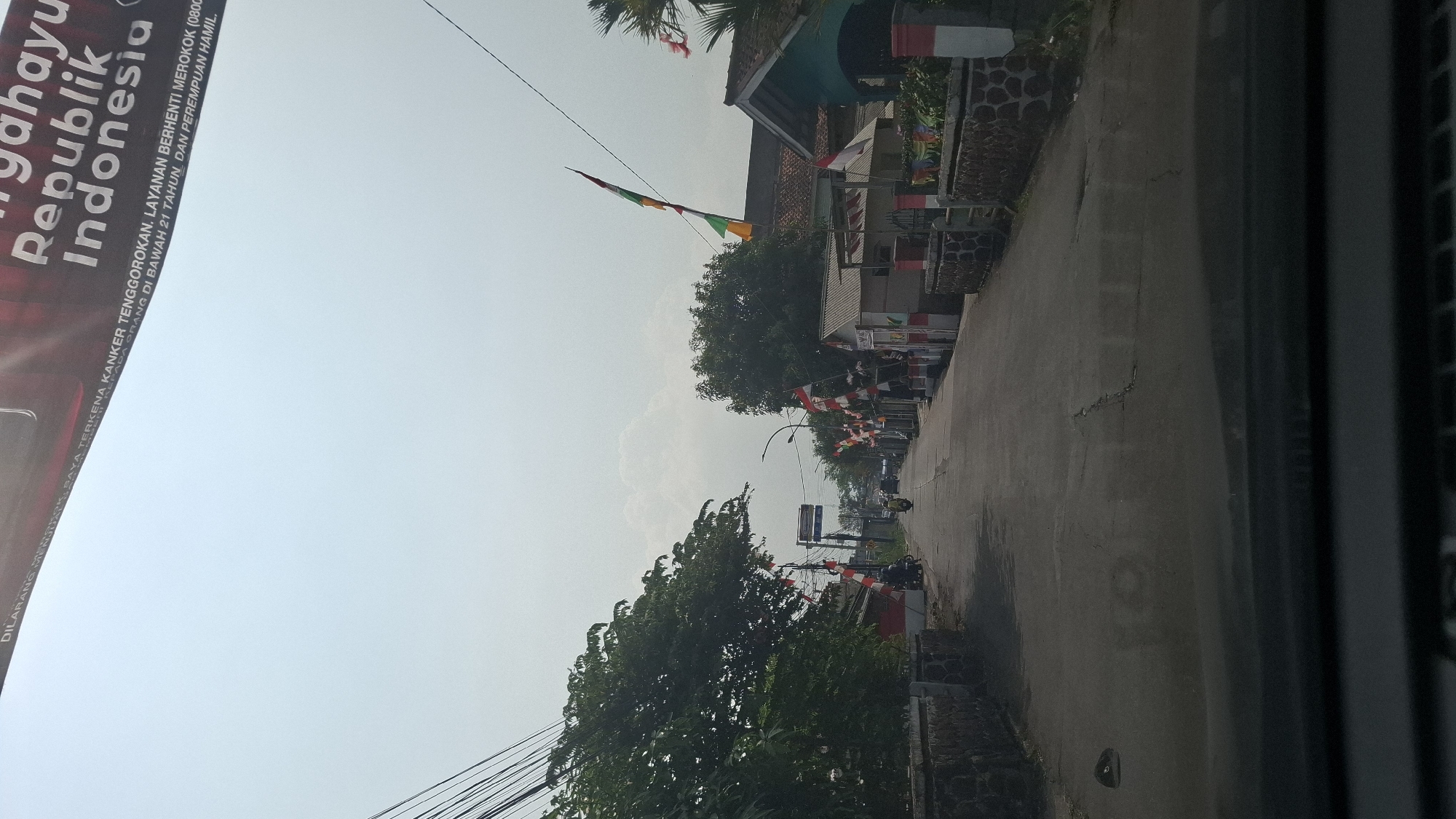 The image depicts a street scene in Warungkondang, Indonesia, decorated with numerous Indonesian flags and festive banners. The flags are predominantly red and white, the national colors, and are displayed on poles along the sides of the road and on buildings. The banners, some of which are partially visible and appear to be anti-smoking or public health messages, hang overhead. The road itself is paved and appears to be a local street, with houses and trees lining either side. A few motorbikes are visible on the road, suggesting ongoing activity. The sky is bright and somewhat hazy, indicating a daytime scene. The overall impression is one of a community celebrating or participating in a patriotic event.