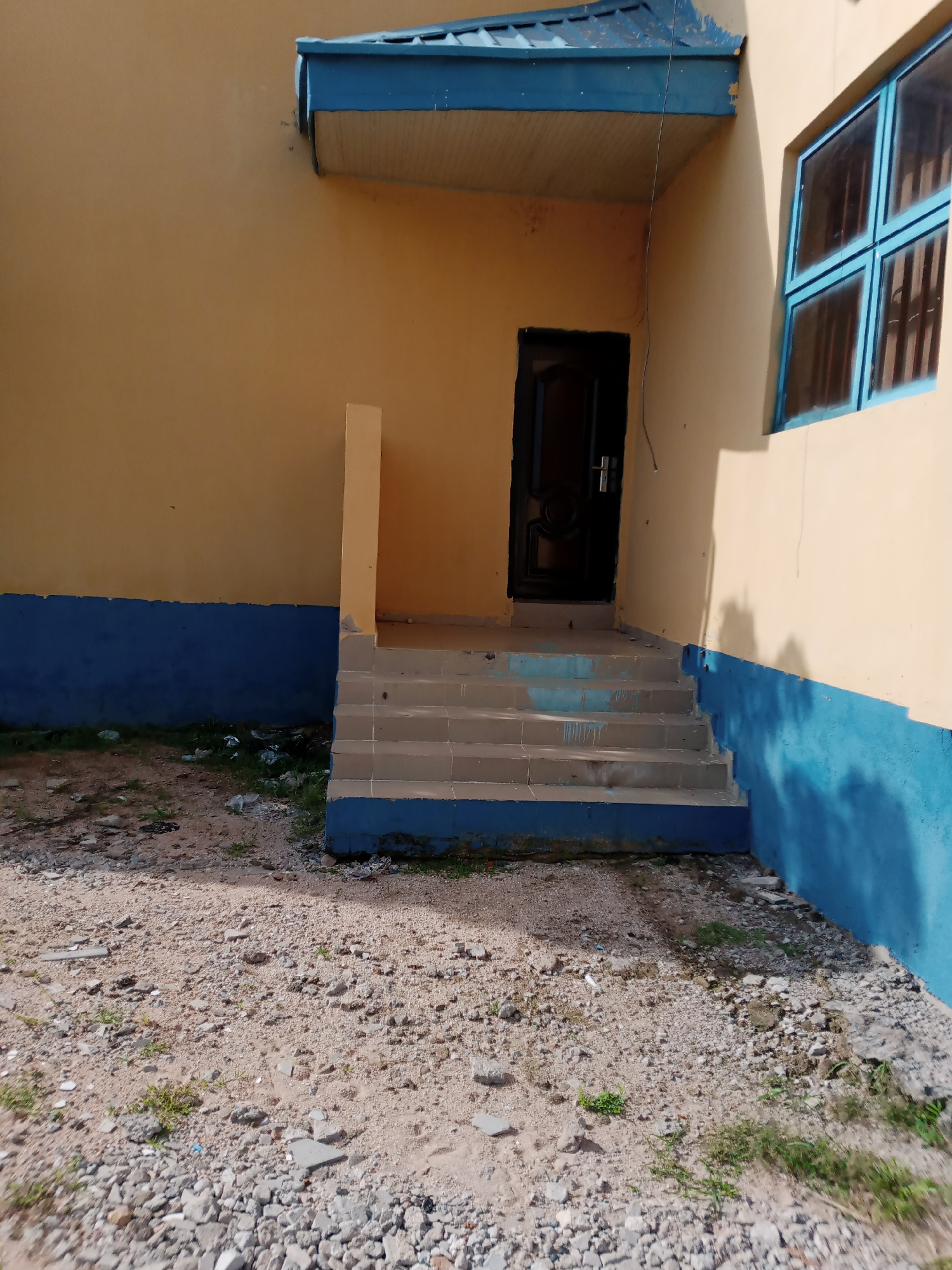 The image depicts an exterior view of a building with yellow walls, accented by a blue trim along the base and a blue overhang above the entrance. A set of five concrete steps leads up to a dark-colored door. To the right of the door, a window with blue framing and multiple panes is visible. The ground in front of the building is unpaved, covered in gravel, sand, and scattered debris, including some green vegetation. The scene appears to be located in an outdoor environment, possibly a public or institutional space. The overall impression is of a somewhat neglected but functional building.