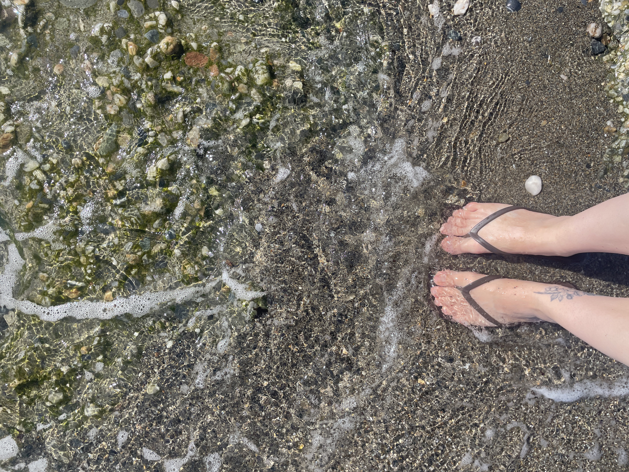 A person's feet, wearing brown flip-flops, are submerged in shallow, clear water at the edge of a beach. The water ripples over a mix of dark sand and small, multi-colored pebbles, some of which are covered in green algae. The sun reflects brightly off the water's surface, creating a shimmering effect. A white foam line from a receding wave is visible above the feet, and more pebbles and some seaweed are scattered on the wet sand further up the beach. The person's left leg has a tattoo of a floral design on the ankle. The location is described as Unknown, Spain.