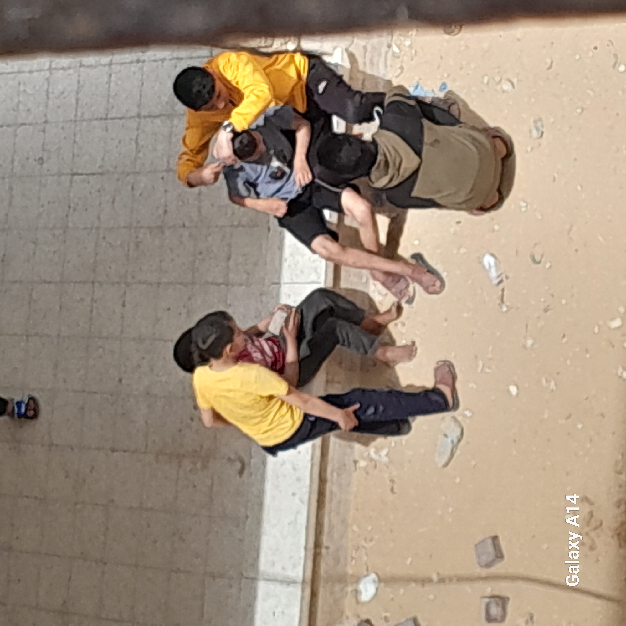 An overhead shot shows several males outdoors on a partially paved and sandy surface. The image is divided by a light-colored concrete or stone curb, with grey square paving stones on the left and light brown sandy or dirt ground on the right.
In the upper-center, a group of three males is interacting. One male, wearing a yellow shirt and a black cap, is positioned over another male in a grey/blue polo shirt and black shorts, appearing to hold him. A third male, dressed in a brown hoodie/jacket and dark pants, stands or kneels close by, observing or participating. Some footwear, including flip-flops, is visible on the ground near this group.
Further down and to the left, another group of two males is present. One male, wearing a red patterned shirt and dark pants, is lying down on the sandy ground. Another male, in a yellow shirt and dark blue pants, is sitting near him. Their posture suggests they are resting or relaxing.
The ground around the individuals has scattered debris, including small white crumpled objects and some dark, square-shaped items. The lighting suggests daylight hours. The image quality is somewhat grainy, possibly due to digital zoom or low light conditions. The text "Galaxy A14" is visible in the bottom right corner.