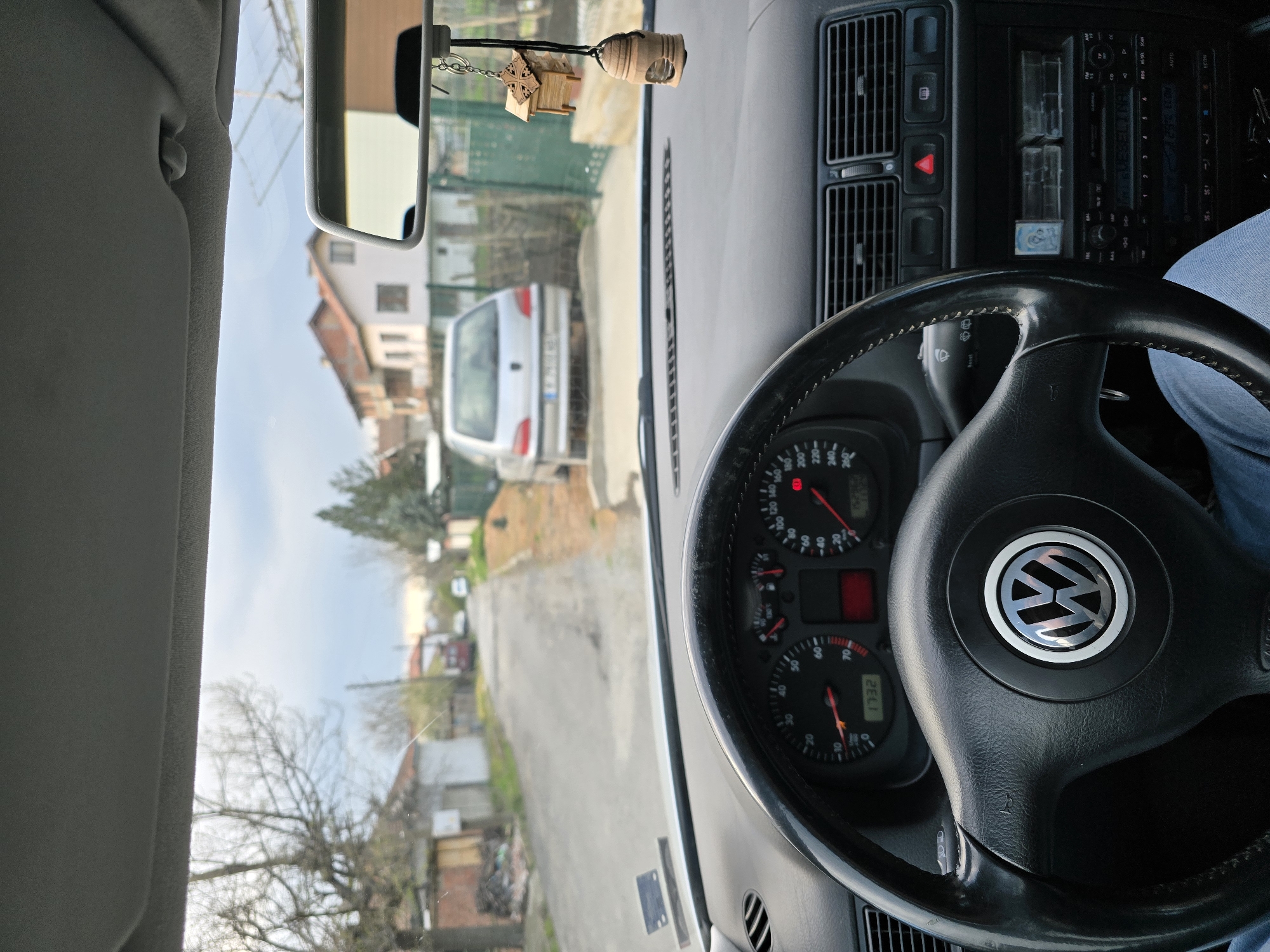 From the driver's seat of a Volkswagen, looking out onto a street in what appears to be Stara Reka, Bulgaria. The car's speedometer reads 1732. A silver car is parked ahead. The location is characterized by residential buildings, trees, and a narrow road. The steering wheel is prominently featured, with the Volkswagen logo clearly visible. The driver's leg is partially seen in the lower right. A wooden ornament hangs from the rearview mirror.