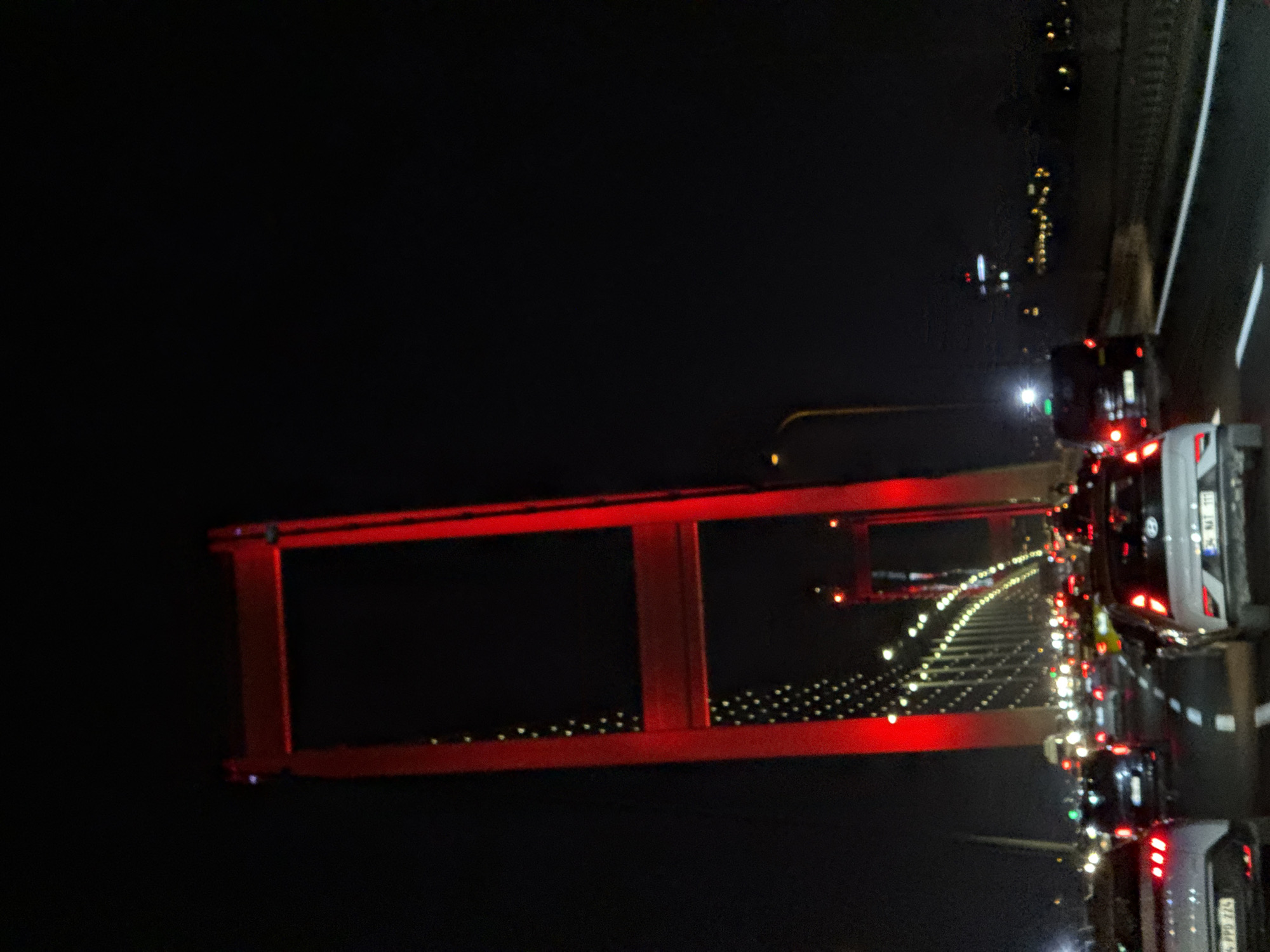 The image captures a nighttime scene on a bridge, likely the Bosphorus Bridge, in Beşiktaş, Turkey. A large, red suspension bridge tower dominates the left side of the frame, illuminated by vibrant red lights. Below and to the right of the tower, the bridge's roadway is visible, filled with a significant amount of traffic. Cars are stopped or moving slowly in multiple lanes, their headlights and taillights creating streaks of light in the darkness. The sky is completely black, suggesting a late hour. In the background, distant city lights are faintly visible, hinting at the urban landscape surrounding the bridge. The overall impression is one of a busy, illuminated infrastructure at night, characteristic of a major metropolitan area like Istanbul.