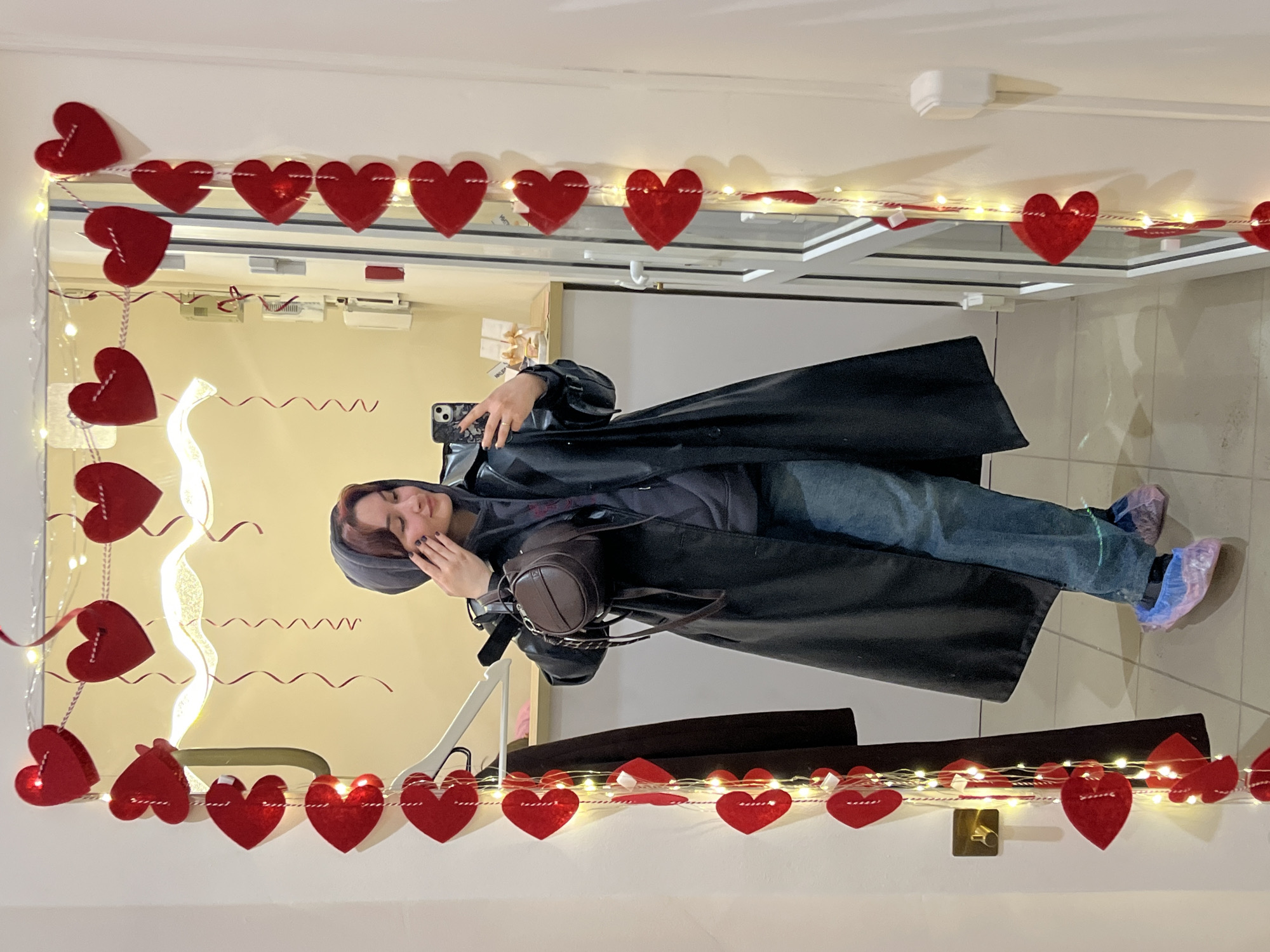 A person stands indoors, positioned in front of a mirror that is decorated with a string of red hearts and small fairy lights. The person is wearing a long, dark coat, jeans, and appears to have booties over their shoes. They are holding a cell phone and seem to be taking a picture of themselves in the mirror. The room appears to be indoors, with a warm light illuminating the background. The photo was taken in Neftekamsk, Russia.