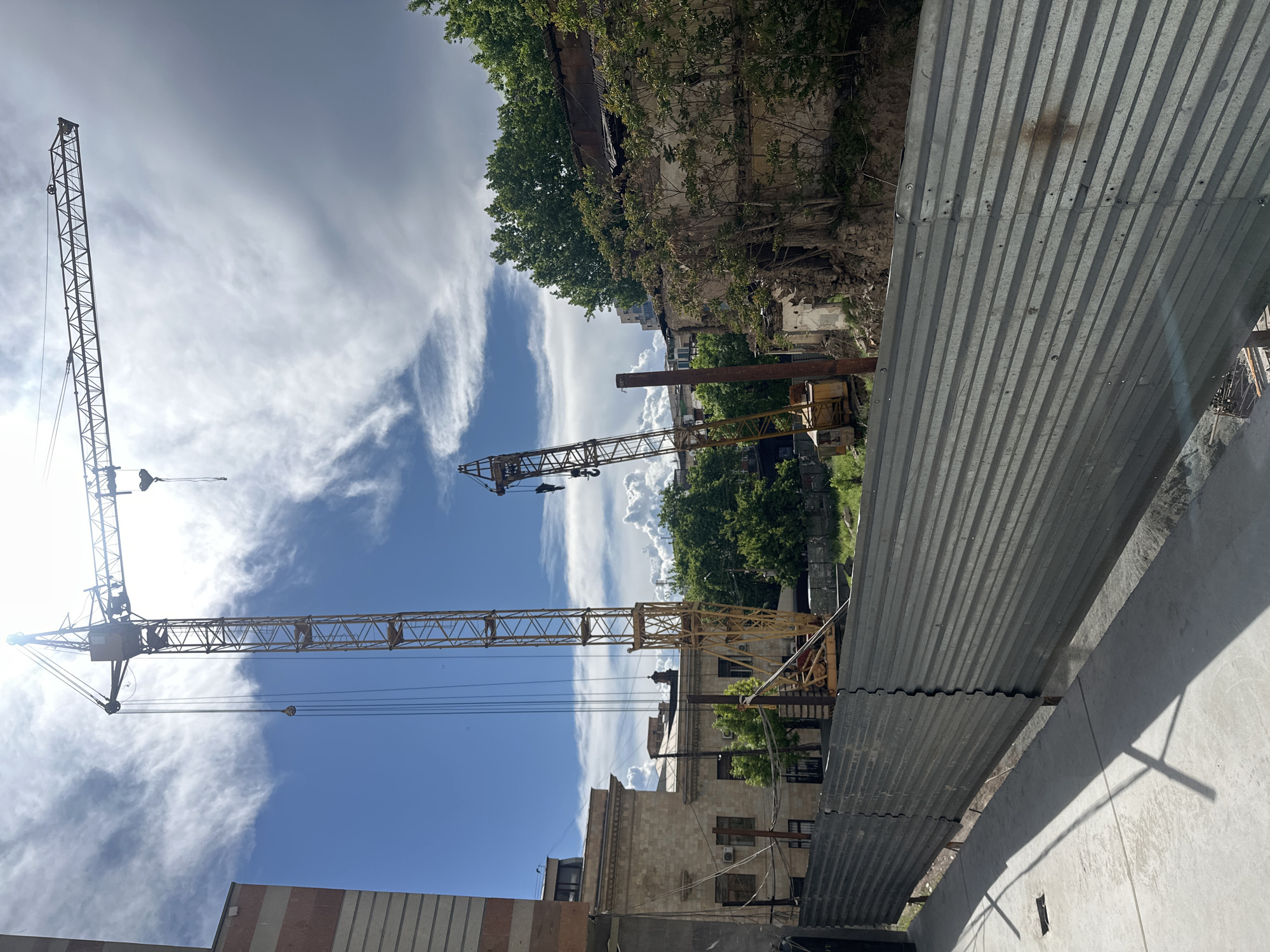 The image captures a scene in Yerevan, Armenia, dominated by construction activity. In the foreground, corrugated metal sheets form a barrier, likely part of a construction site enclosure. Behind the barrier, there are multiple construction cranes of varying sizes, positioned against a backdrop of blue sky with scattered clouds. The cranes are of a lattice-type construction.
Adjacent to the construction site is an older building partially covered with greenery, suggesting age and potential disrepair. Some portions of the building appear to be crumbling. Other buildings and trees can be seen in the background, indicating an urban environment. The overall atmosphere suggests urban development juxtaposed with elements of the older city.