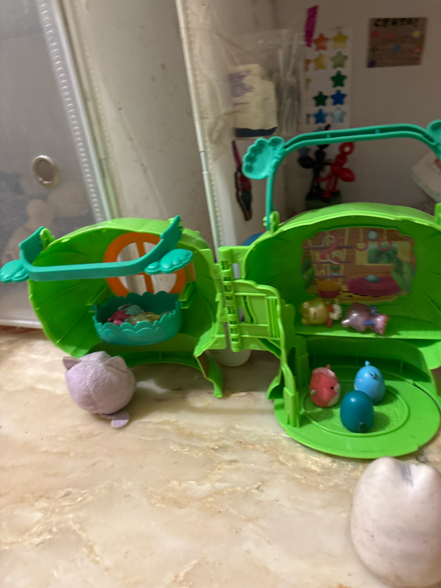 The image shows a green, leaf-shaped toy playset. The playset is open, revealing different compartments and miniature figures. In the left part of the playset, there is a green basket with small, colorful items inside, and a purple plush toy sits next to it. The right side of the playset contains shelves with small toy figures resembling animals, and a backdrop depicting a room interior. In front of the playset, there are three small, brightly colored toy figures on a green platform, with another similar figure slightly out of focus in the foreground to the right. The playset rests on a marble-like surface. The background is blurred and appears to be an indoor setting with some shelving and decorative items. No specific location in Ashvem, India is visible or implied in the image.