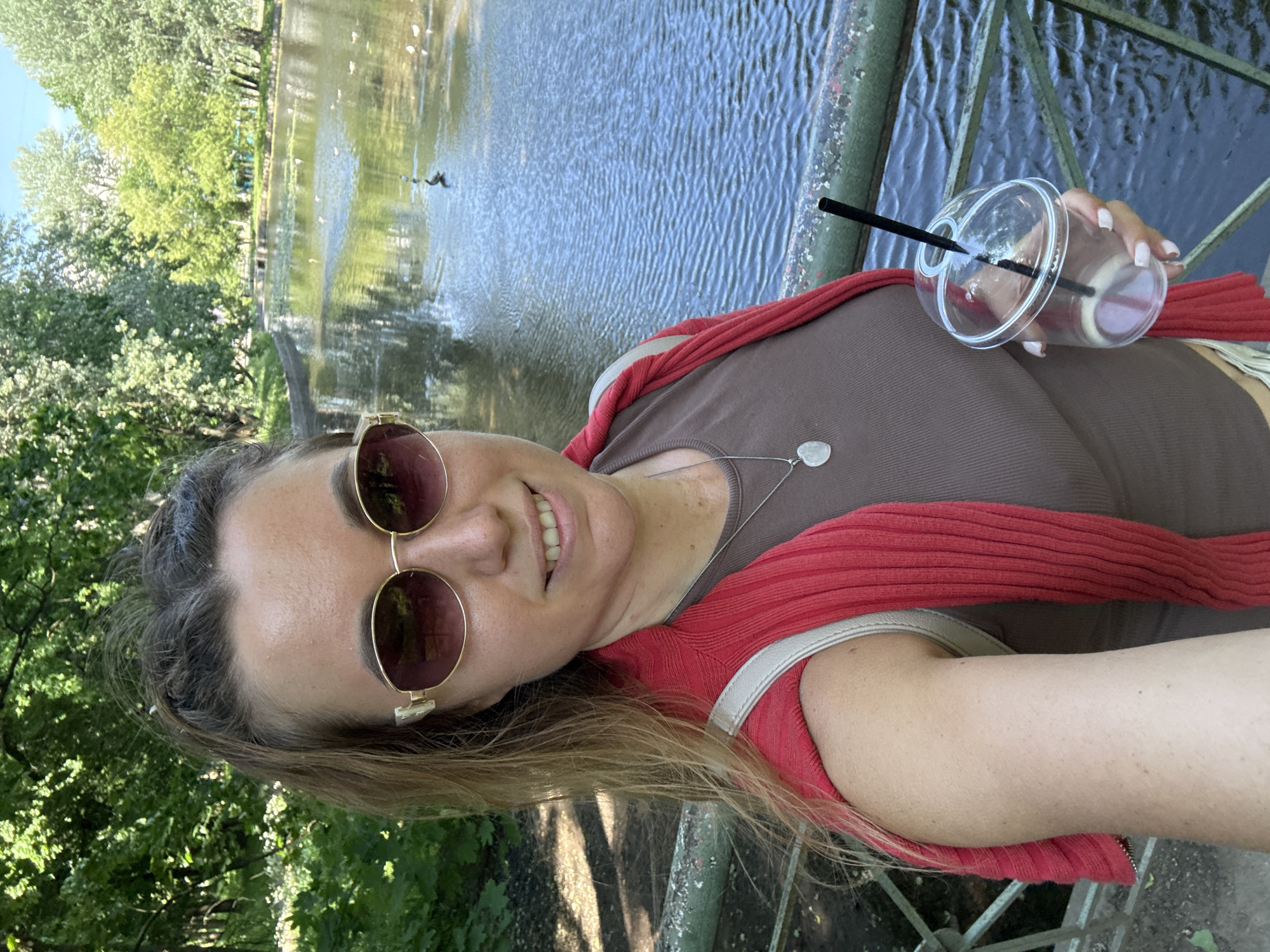A woman is taking a selfie in front of a lake. She is wearing sunglasses, a brown tank top, and a red sweater draped over her shoulders. She is holding a clear plastic cup with a black straw in her right hand. The lake is surrounded by trees, and there are several birds on the water. The scene appears to be in a park.