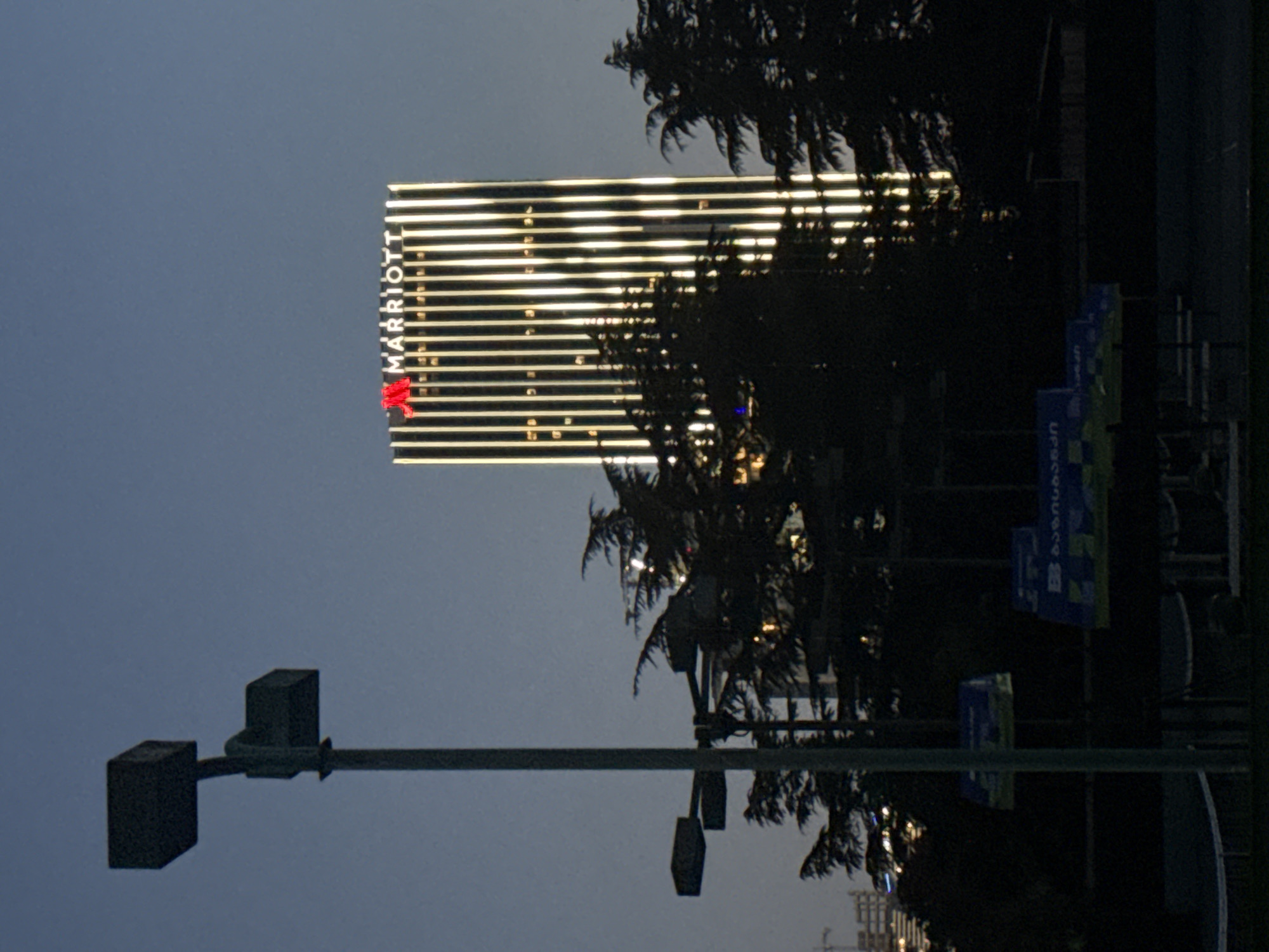 The image depicts the Batumi Marriott Hotel in Batumi, Georgia, at dusk or dawn. The hotel, a high-rise structure, is illuminated with horizontal lines of light. A Marriott logo, featuring a distinctive emblem, is visible on the building. A light standard with two lights is in the foreground. Trees and possibly vegetation partially obscure the hotel's lower portion. There are vertical banner advertisements also in the foreground. The sky is a gradient of dark blue to light gray.