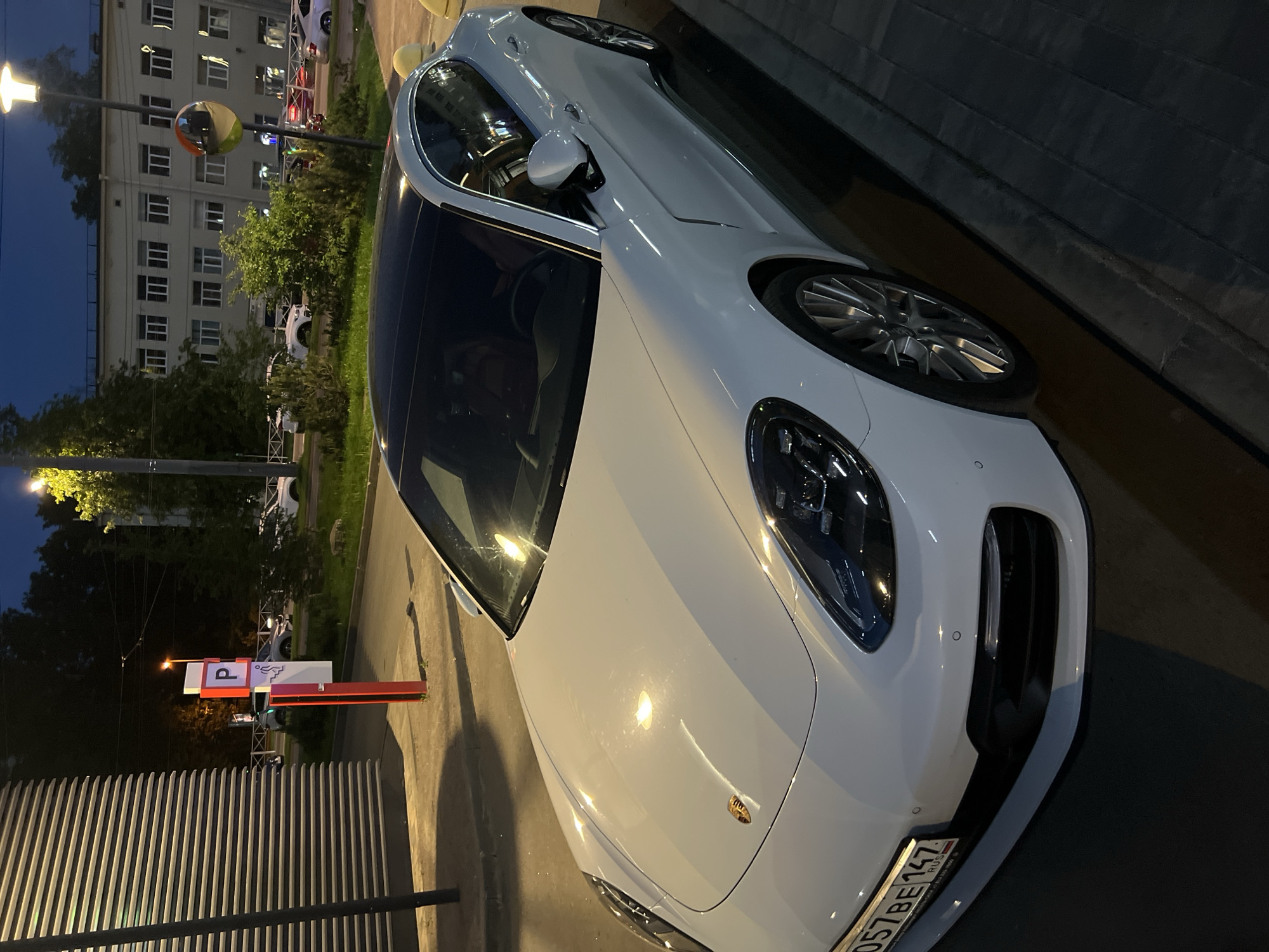 A white Porsche Panamera is parked on a city street at dusk. The car is the main subject, positioned at an angle with its front left side facing the camera. It has a black roof and silver rims. The car's license plate, partially visible, reads "057BE 147 RUS".
In the background, a multi-story building with multiple windows is visible, as is a lamppost with a yellow light. A parking sign with a red pole and a white sign displaying a "P" symbol and a silhouette of a person is also present. Trees and other parked cars can be seen further in the background. The ground is paved with bricks, and there is a sidewalk. The lighting suggests it is nighttime or late evening.