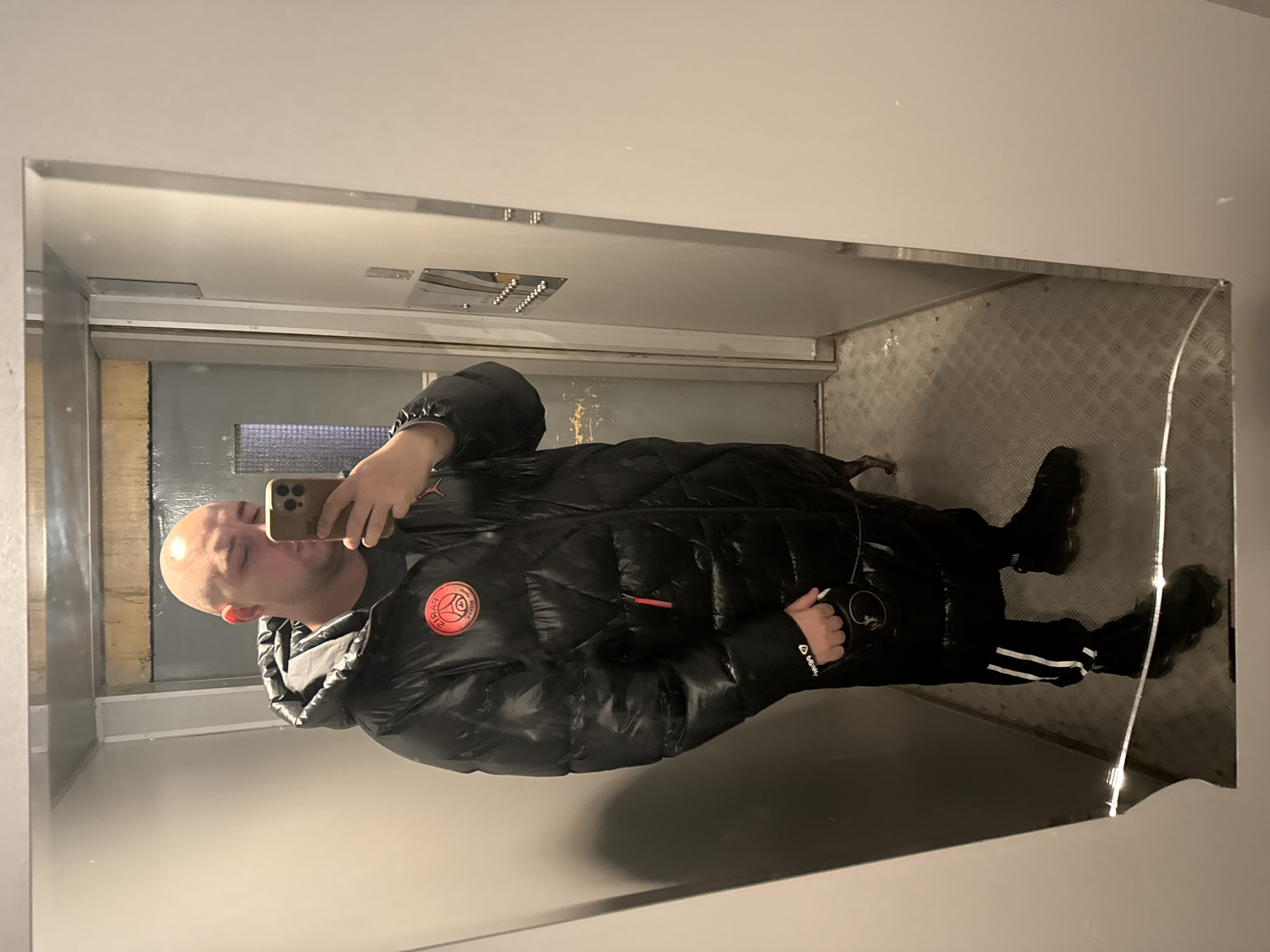 A bald man in a black puffer jacket with a PSG logo is taking a selfie in a mirror inside what appears to be an elevator. The elevator's interior has a metal floor with a diamond plate pattern and a gray wall. The man is wearing black pants with white stripes. His phone is a light brown color. The location in Pleven, Bulgaria, is not discernible from the image.