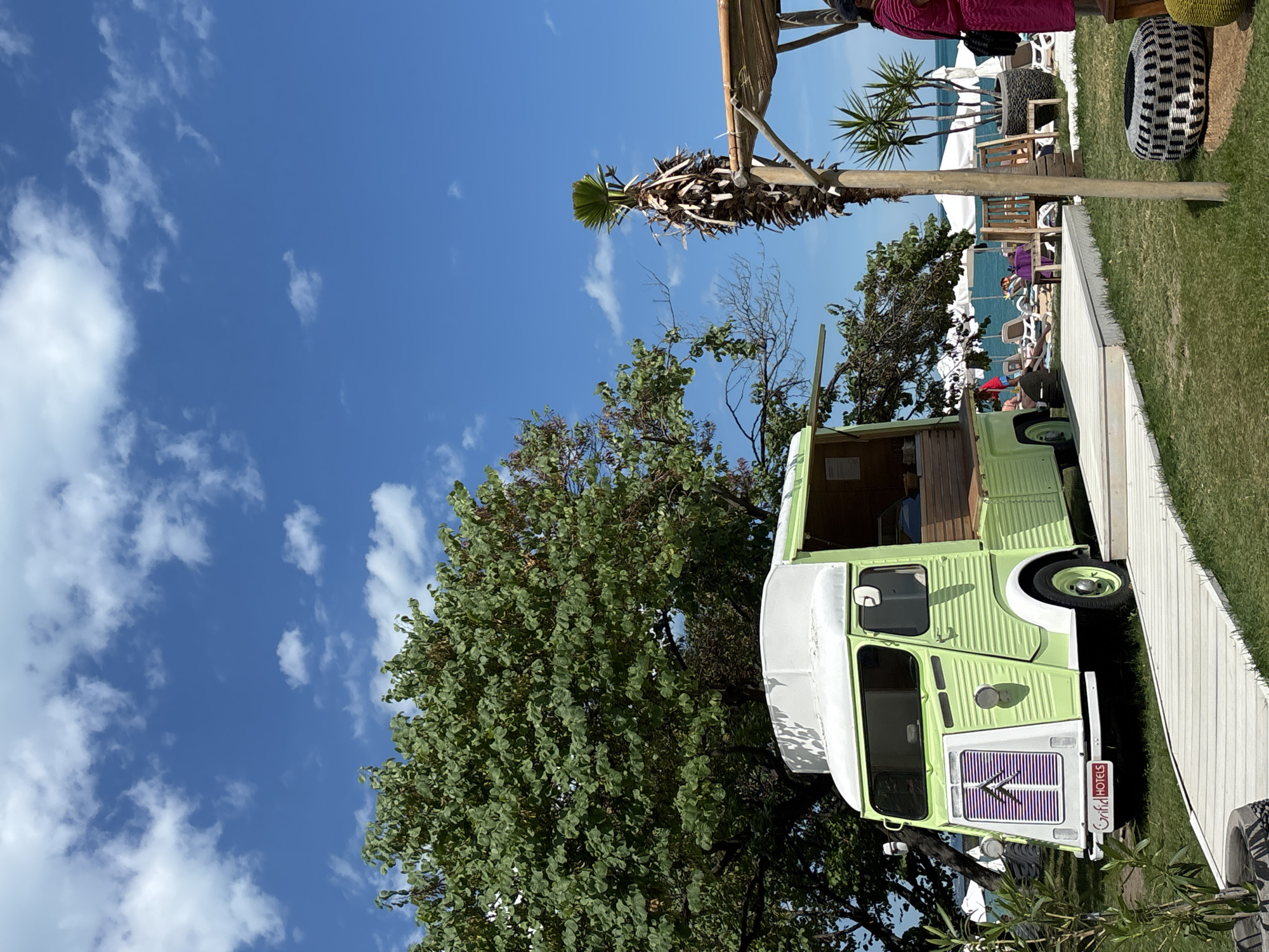 A light green Citroen H Van, converted into a food truck, is parked on a grassy area with a wooden path leading to it. The van has a white roof and a distinctive front grille with purple stripes. A small sign with "GRIFID HOTELS" is visible on the front bumper. The van is located outdoors, possibly at a beach resort, given the presence of beach chairs, umbrellas, and the sea in the background. A palm tree and other greenery are also present in the scene. The sky is blue with scattered white clouds. A person in a pink garment is partially visible on the right side of the image, near a rustic wooden structure with a thatched roof. The overall setting suggests a sunny day at a leisure location. The location is identified as Varna, Bulgaria.