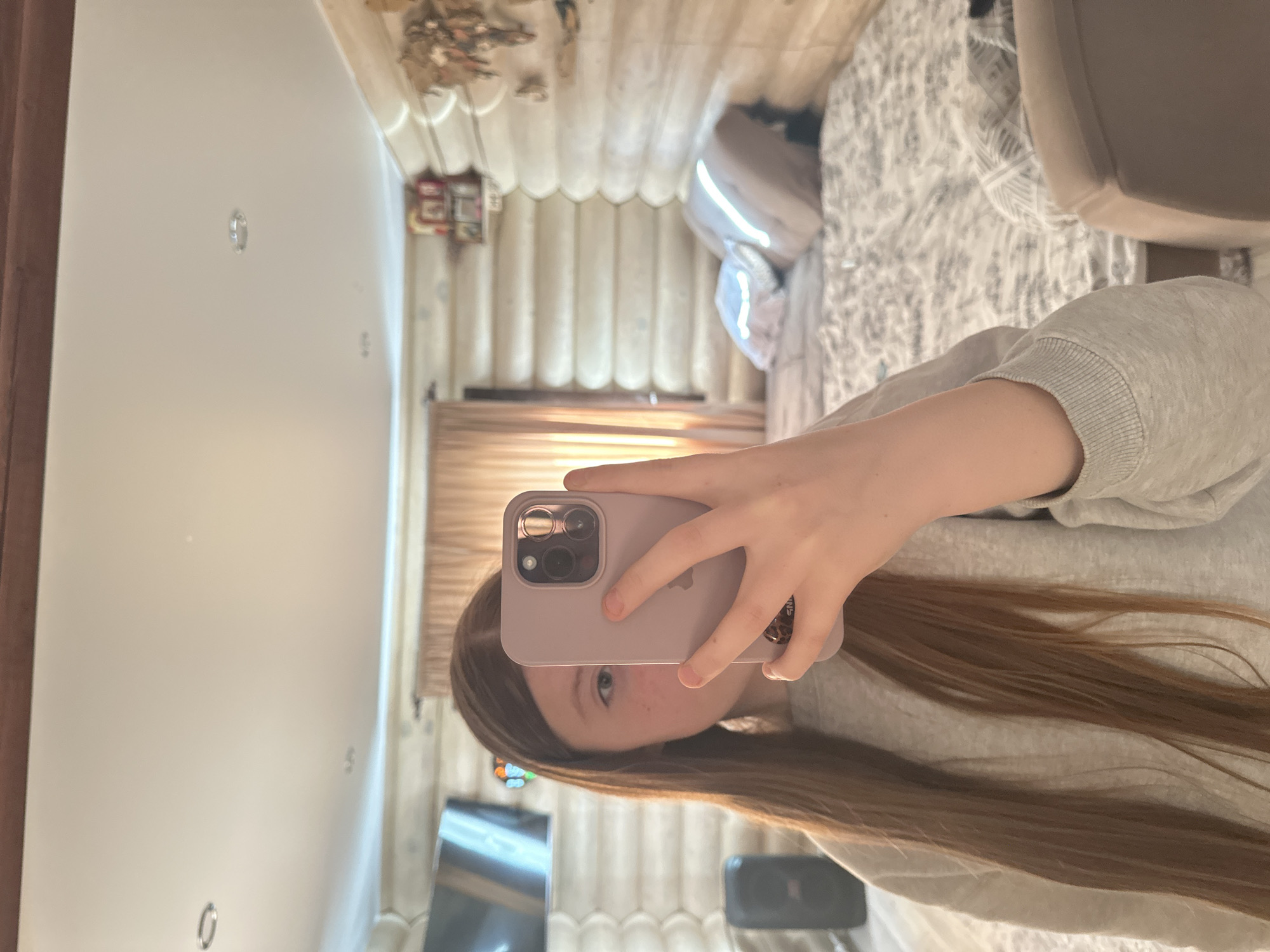 A girl is taking a selfie in front of a mirror. She is holding a lilac phone in her right hand. The girl has long brown hair and is wearing a gray sweatshirt. In the reflection, a log cabin-style interior is visible, with a bed, chair, television, and assorted objects. The location is possibly in the city of Olkhovka, Russia.