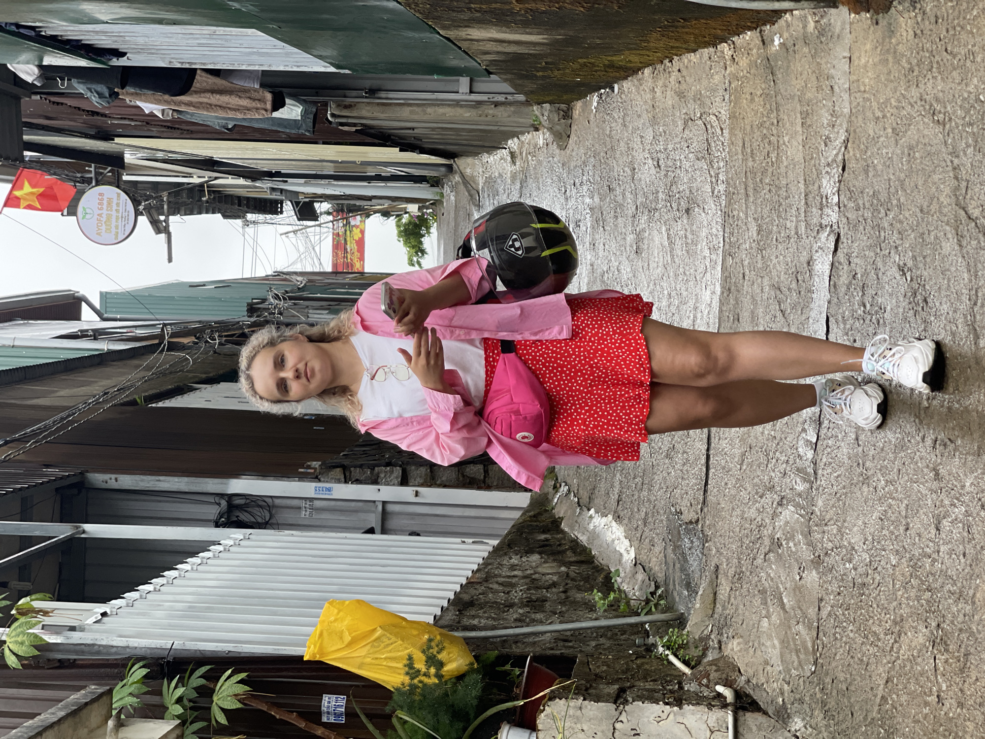 A woman is standing in a narrow alley in Lang Biang - Đà Lạt, Vietnam. She is wearing a white t-shirt, a pink shirt, a red skirt with white polka dots, and white sneakers. She has a black motorcycle helmet and a pink fanny pack. She is holding a phone in her hand. The alley is lined with buildings made of corrugated metal and stone. There is a Vietnamese flag visible. The ground is wet and appears to be concrete.