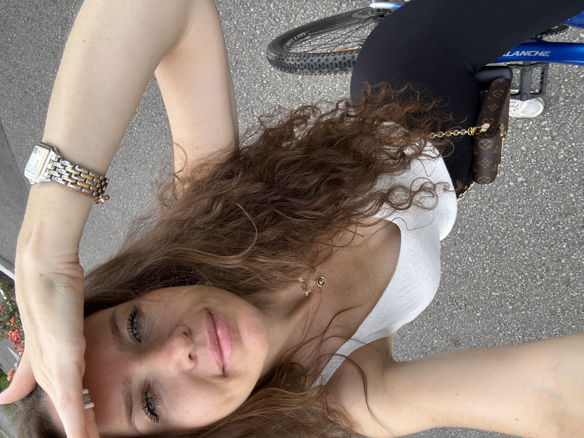 A woman is taking a selfie from a high angle, looking at the camera with her right hand shielding her eyes from the sun. She has long, wavy brown hair and is wearing a white tank top, black leggings, and a small designer purse with a gold chain. She is also wearing a watch on her left wrist. The background appears to be a paved surface, likely a road or sidewalk, with some greenery and flowers visible in the upper left corner. The image does not provide any specific clues to determine its location in Moscow, Russia.