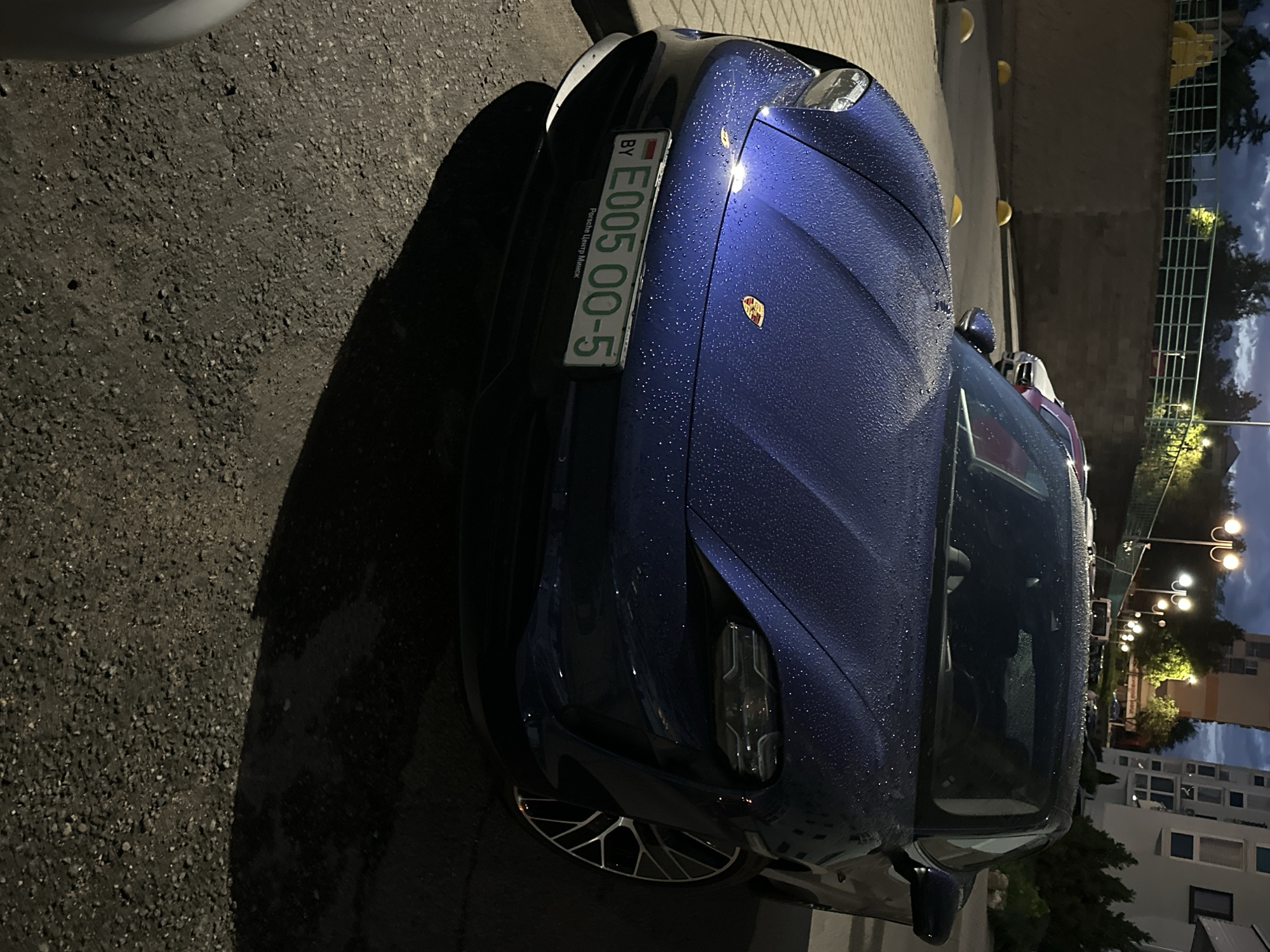 A blue Porsche Taycan is parked on the side of a street in Minsk, Belarus. The license plate reads "E005 00-5" and includes the "BY" country code. The car is wet, likely from rain, with water droplets visible on the hood. The surrounding environment includes a paved street, a building, and streetlights. The lighting suggests it is either dusk or early evening.