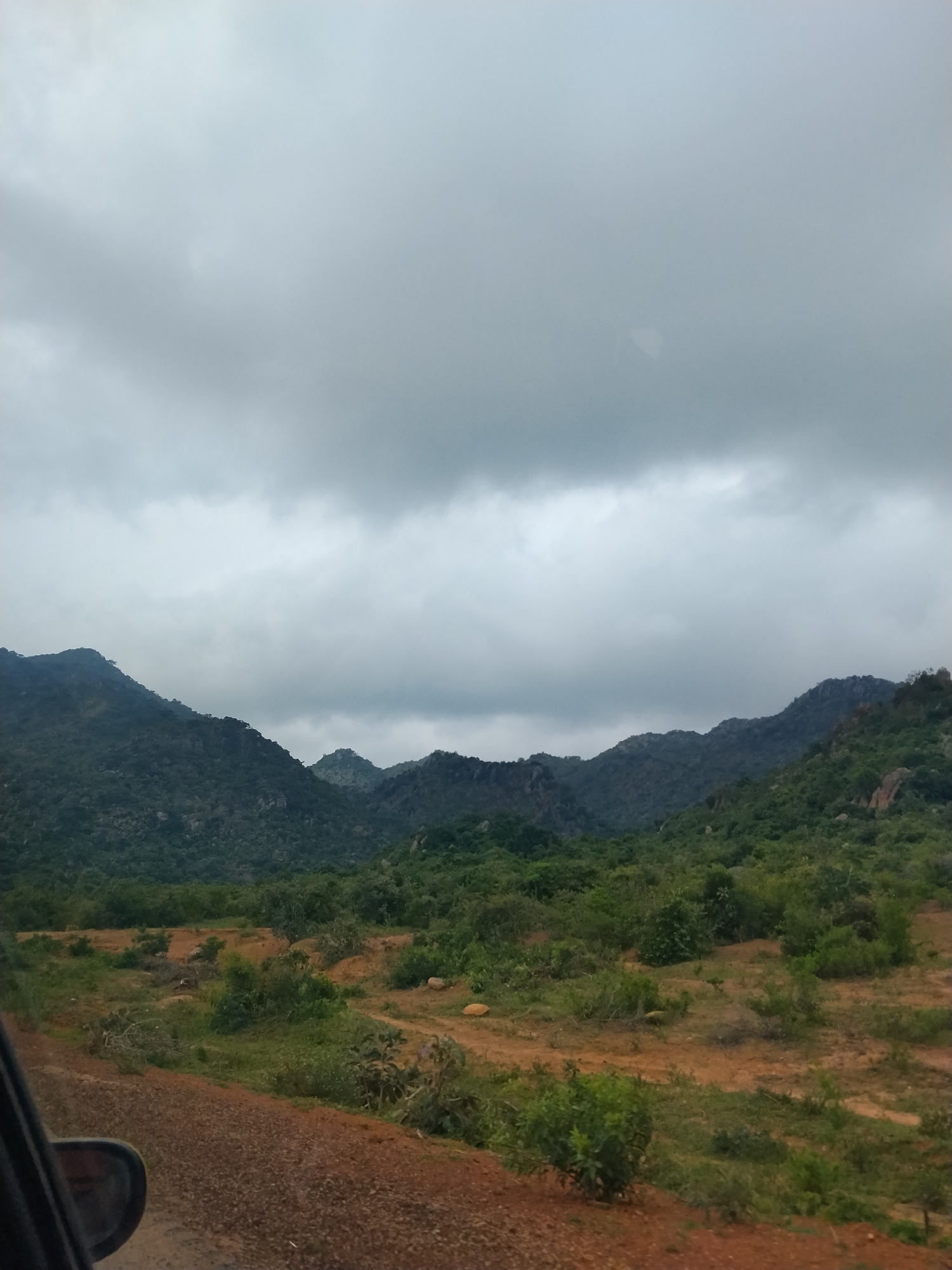 The image shows a landscape scene featuring rolling, forested hills under a cloudy sky. In the foreground, a dirt road with a car's side mirror is visible on the left. The terrain is predominantly brown earth with scattered green vegetation. The hills are covered in dense green trees and shrubs. The sky is overcast with heavy, gray clouds. The scene appears to be a natural, rural environment, consistent with the geography of Dorowa, Nigeria, known for its hilly terrain and vegetation. There are no people or specific actions depicted in the image.