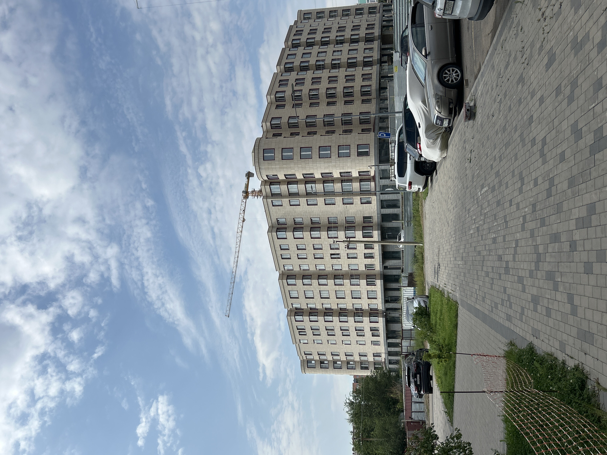 In Astana, Kazakhstan, a modern, multi-story apartment building with a light-colored facade and numerous windows is visible under a sky with scattered white clouds. A construction crane extends diagonally across the upper portion of the frame, indicating ongoing development. The foreground features a paved walkway and a parking lot with several cars, including a dark SUV, a silver sedan covered with a tarp, and a light-colored SUV. A temporary orange safety fence encloses a grassy area adjacent to the walkway. In the background, trees and fencing are also present.