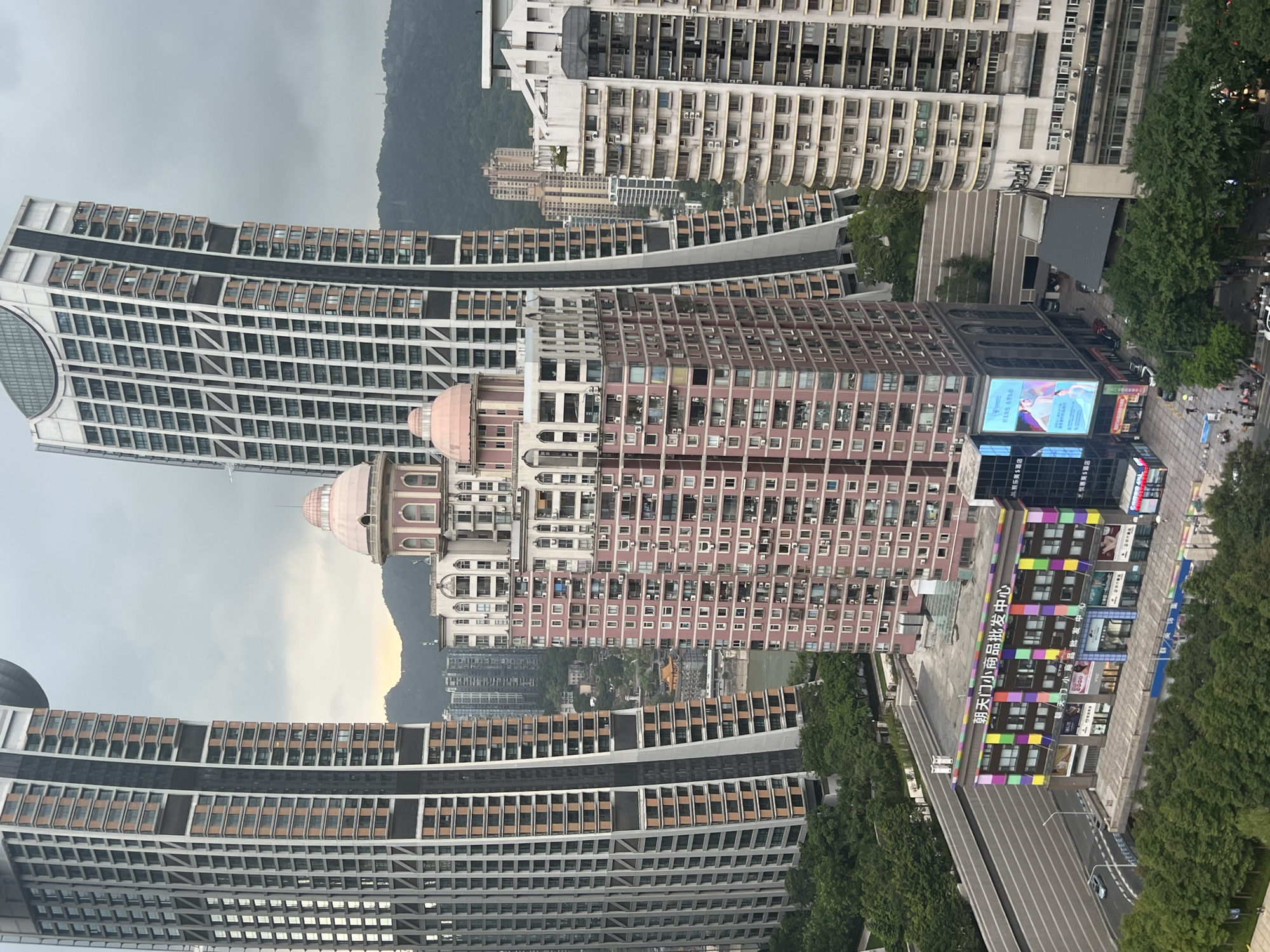 This is a high-angle, full shot of a cityscape in Yuzhong District, China, likely taken from a high-rise building. The scene is dominated by a cluster of tall, modern buildings with distinct architectural styles.
In the foreground, a large, pink, multi-story building stands prominently. It features numerous balconies with air conditioning units and is topped with two distinctive pink domes. To its left and right are two massive, sleek skyscrapers with angular designs and numerous windows, one on the left with a curved, metallic top, and the other on the right leaning slightly inwards. Further in the distance, a dense urban landscape unfolds with more high-rise buildings of varying heights and designs, some appearing older than the foreground structures. A river is visible between some of the buildings, and green hills form the backdrop.
At the base of the pink building, a colorful commercial structure with a vibrant, striped facade is visible. Various storefronts and a large digital billboard are present on this lower building. Trees line the streets and surround the base of some of the buildings, indicating some level of urban greenery.
A few cars are visible on the road in front of the commercial building, and several pedestrians can be seen walking on the sidewalks, though they are too small to discern any specific details. The sky is overcast, suggesting a cloudy or potentially rainy day. The overall impression is one of a dense, modern, and vertically oriented urban environment.