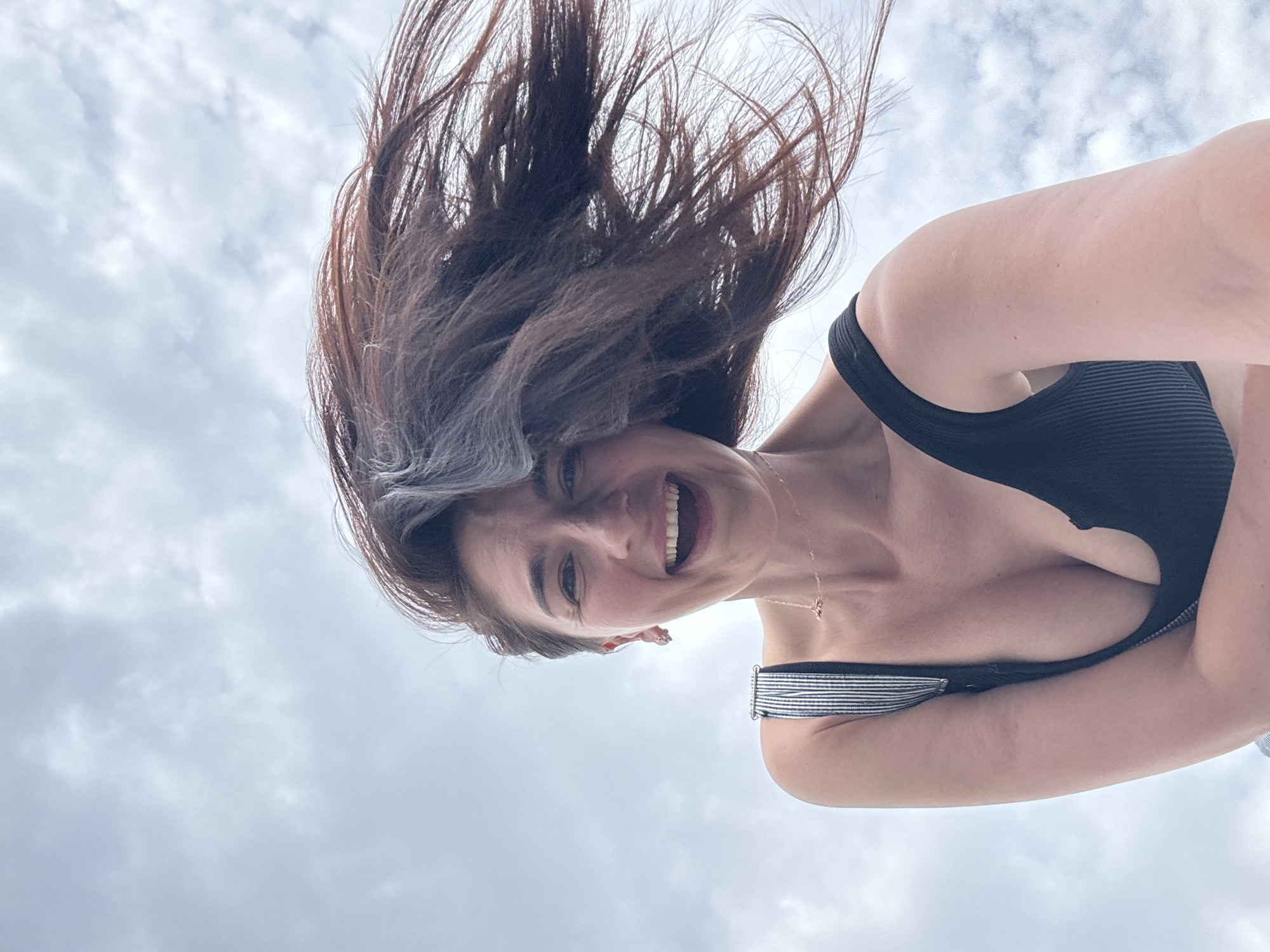 A woman with dark brown hair and fair skin smiles at the camera. She is wearing a black bathing suit top. Her hair is tossed around. The sky is overcast with clouds in the background. There is no other information available about the location, particularly whether it is in Sochi, Russia.