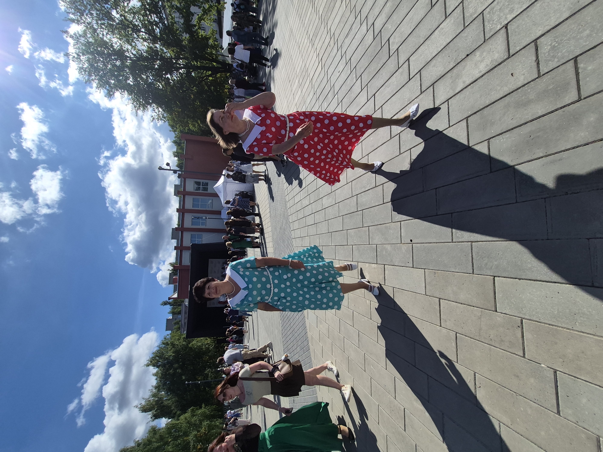 This is a medium shot of a sunny outdoor scene with several people walking on a paved area. Two women in the foreground are prominently featured, both wearing retro-style polka dot dresses. The woman on the right wears a red and white polka dot dress with a white collar and belt, and white shoes. The woman to her left wears a teal and white polka dot dress with a white collar and belt, and white shoes. Both women appear to be walking towards the left side of the frame. In the background, there is a crowd of people, a stage, tents, trees, and buildings. The sky is blue with scattered white clouds and the sun is shining brightly, casting long shadows on the ground. The paved area is made of grey rectangular stones. This scene is likely taking place in Chaikovsky, Russia, possibly at a public event or festival.