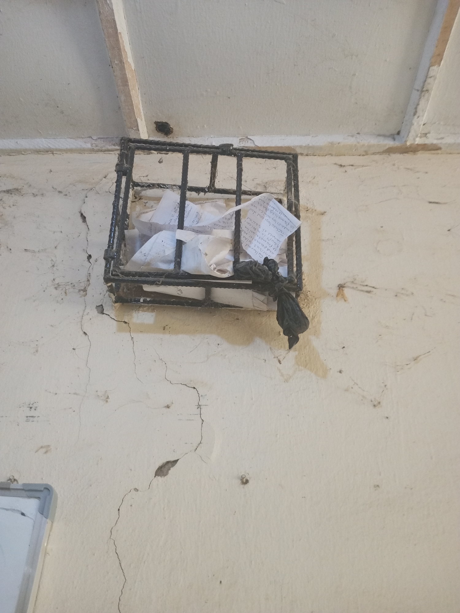 This media file captures a close-up, angled view of a metal cage attached to a cracked, light-colored wall. The cage contains crumpled papers with handwritten text. A black plastic bag is tied to the lower part of the cage. The wall has visible cracks and some staining, suggesting an older structure. The ceiling above appears to be made of wooden planks, some of which are discolored. In the upper left of the image, a dark hole is visible on the ceiling or wall junction. The scene is likely indoors, given the ceiling and wall construction, and the lighting suggests it is daytime.
The visible text on the papers appears to be in English and contains phrases such as "Cerises," "COMM," "Cultural to," "various cultu," "It promote kai," "Djups in africa," "Gannel of social," "and Primotes rosak.CGA," "Technological dev," "in Promoting Race," "means by which wa," and "U reducas the." The text is fragmented and difficult to fully decipher, but it seems to discuss culture, technology, and social aspects, possibly related to development or promotion.
The overall impression is one of a utilitarian or possibly neglected space, with the cage and papers potentially serving as a makeshift bulletin board or storage. The location cues, such as the wall texture and ceiling construction, combined with the given location context of Jalingo, Nigeria, suggest an environment common in that region. There are no people visible in the image, and the focus is on the cage, papers, and the distressed wall.