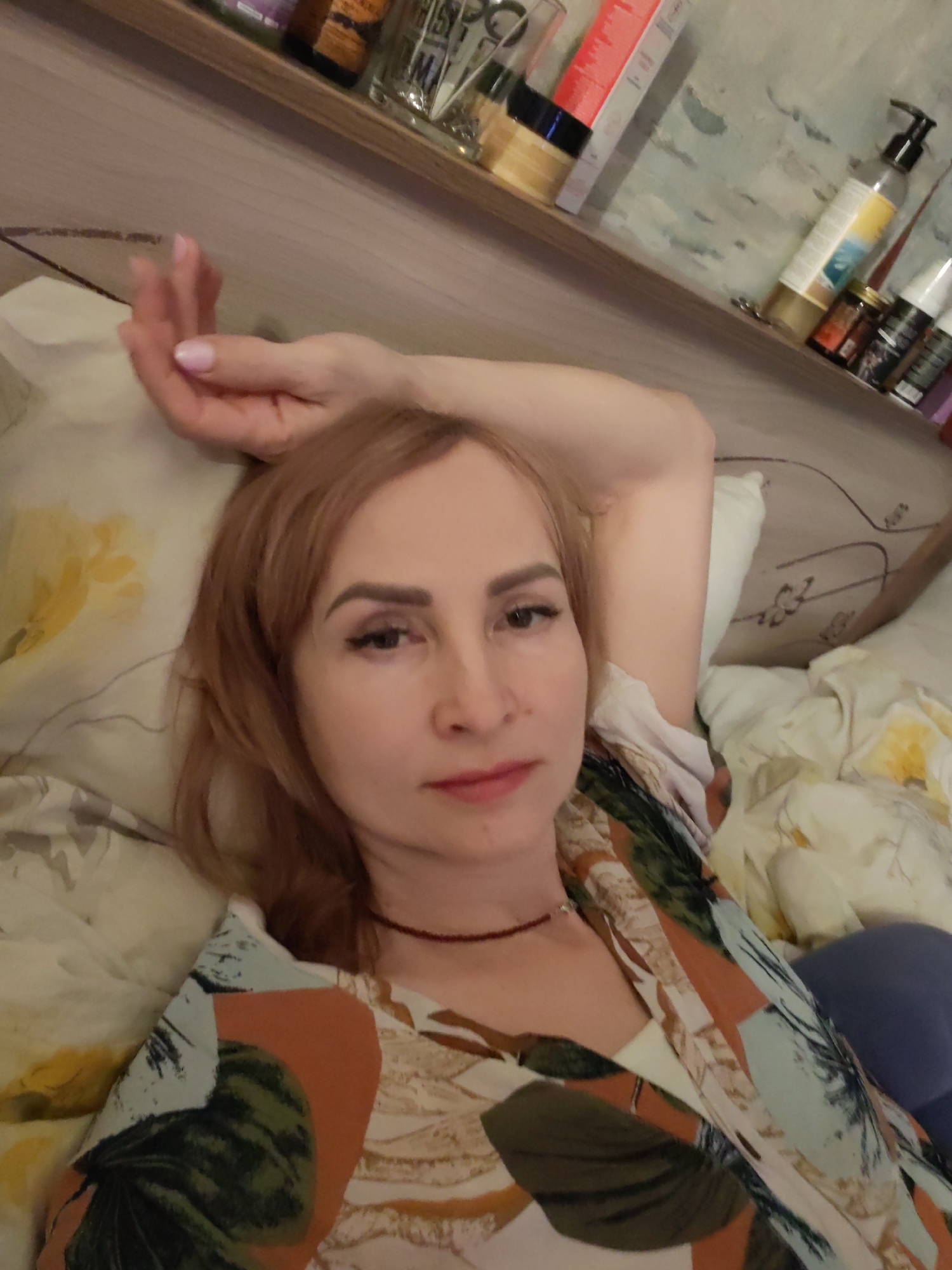 A woman is lying in bed. She has light brown hair and a slight smile. She is wearing a light-colored top with a floral pattern. Her left arm is raised above her head. Behind her is a pillow with a yellow floral pattern. Above her head is a shelf with various bottles and containers. The photograph was taken at an indoor location, possibly a bedroom, in Chaikovsky, Russia.
