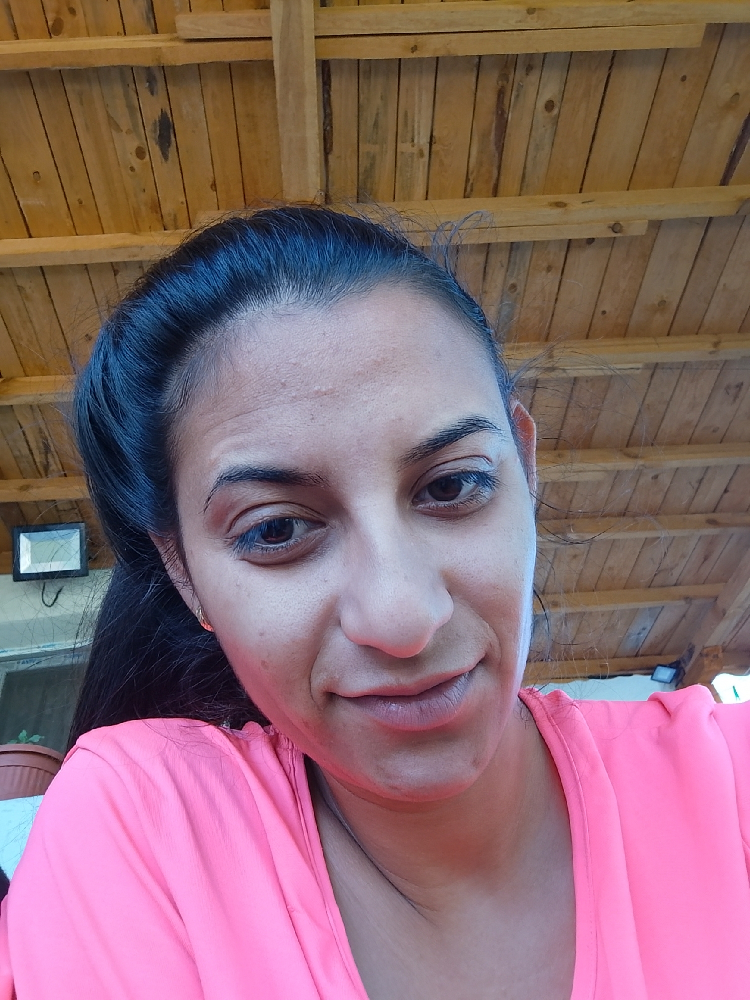 A woman is looking directly at the camera in a close-up shot. She has dark hair pulled back, and her face is mostly visible. She is wearing a bright pink t-shirt. The background shows a wooden ceiling with visible beams and planks, suggesting an outdoor or covered area. To the left of the woman, a small rectangular light fixture is mounted on a wall. A hint of a potted plant and a window can be seen further to the left. The lighting appears to be natural, casting soft shadows on her face. The woman's expression is neutral, with her lips slightly parted. The overall setting suggests a casual, possibly domestic or recreational, environment.