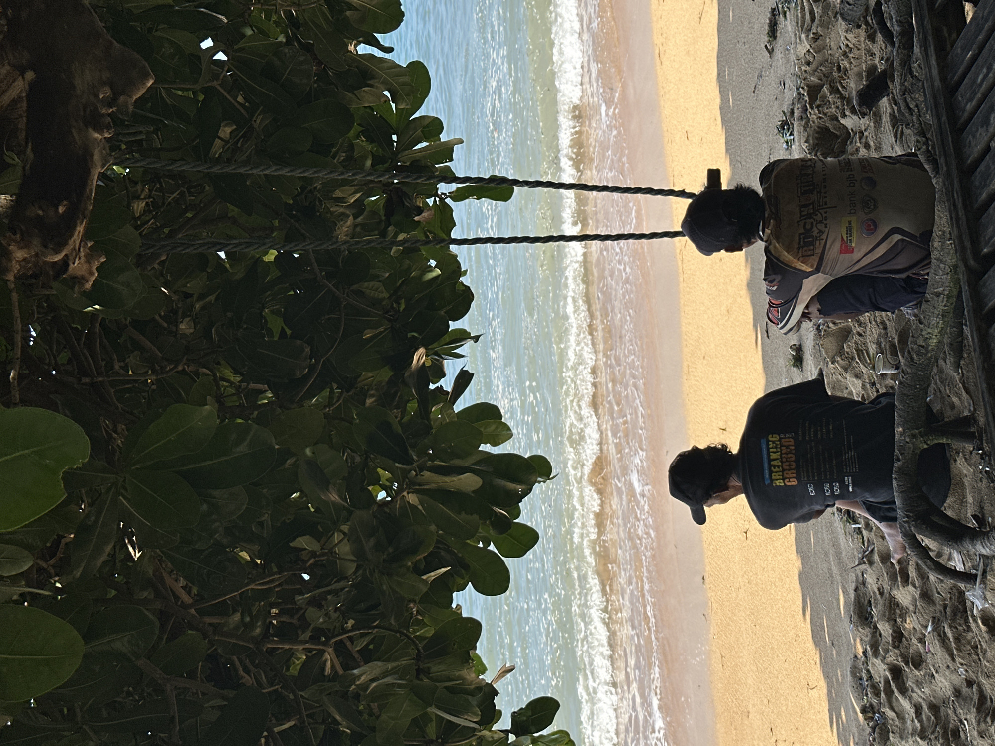 The image depicts a serene beach scene in Denpasar, Indonesia, framed by the lush foliage of a tree. Two individuals, seen from behind, are seated on the sand, their gazes directed towards the tranquil ocean. The person on the left wears a dark baseball cap and a t-shirt with text that appears to be a band or event name, "BREAKING GROUND." The individual on the right, also wearing a cap, is dressed in a patterned jacket with visible text and logos, including "bank bjb". They are seated on a makeshift wooden bench, surrounded by the natural elements of the beach, including sand, driftwood, and scattered debris. The gentle waves of the sea lap at the shore, creating a peaceful ambiance. The lighting suggests it is late afternoon, casting a warm glow on the scene. The presence of a swing hanging from the tree adds a whimsical touch to the otherwise tranquil setting.