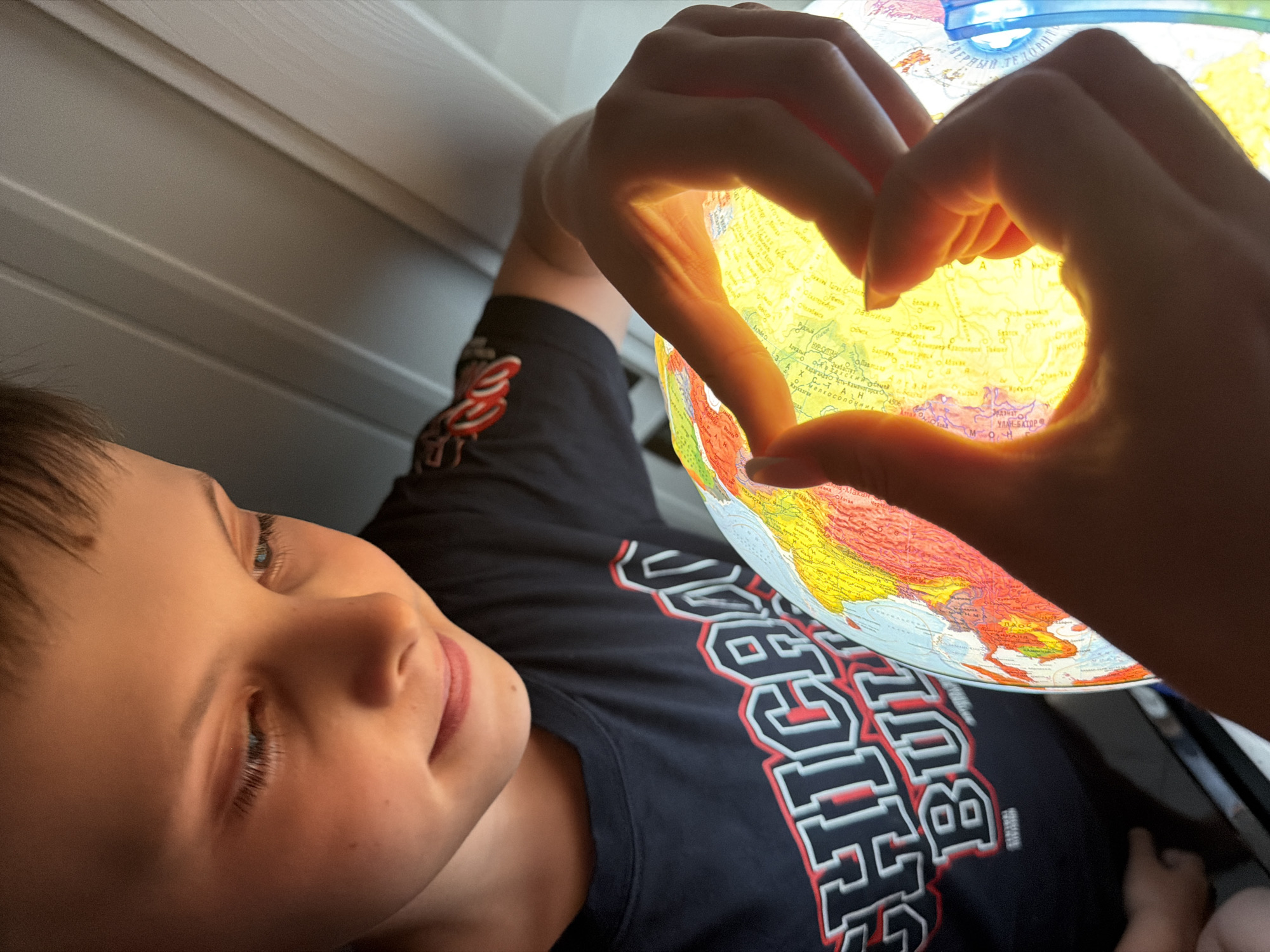 A young boy, wearing a dark t-shirt with white and red lettering, lies on his back. Above him, a pair of hands forms a heart shape around an illuminated globe, focusing on the Asian continent. The globe is positioned between the viewer and the boy. Behind the boy, a wall with horizontal panels is visible. The location is unknown. The boy's expression is peaceful. The focus of the scene appears to be on geography and learning, highlighted by the heart shape symbolizing love for the world.