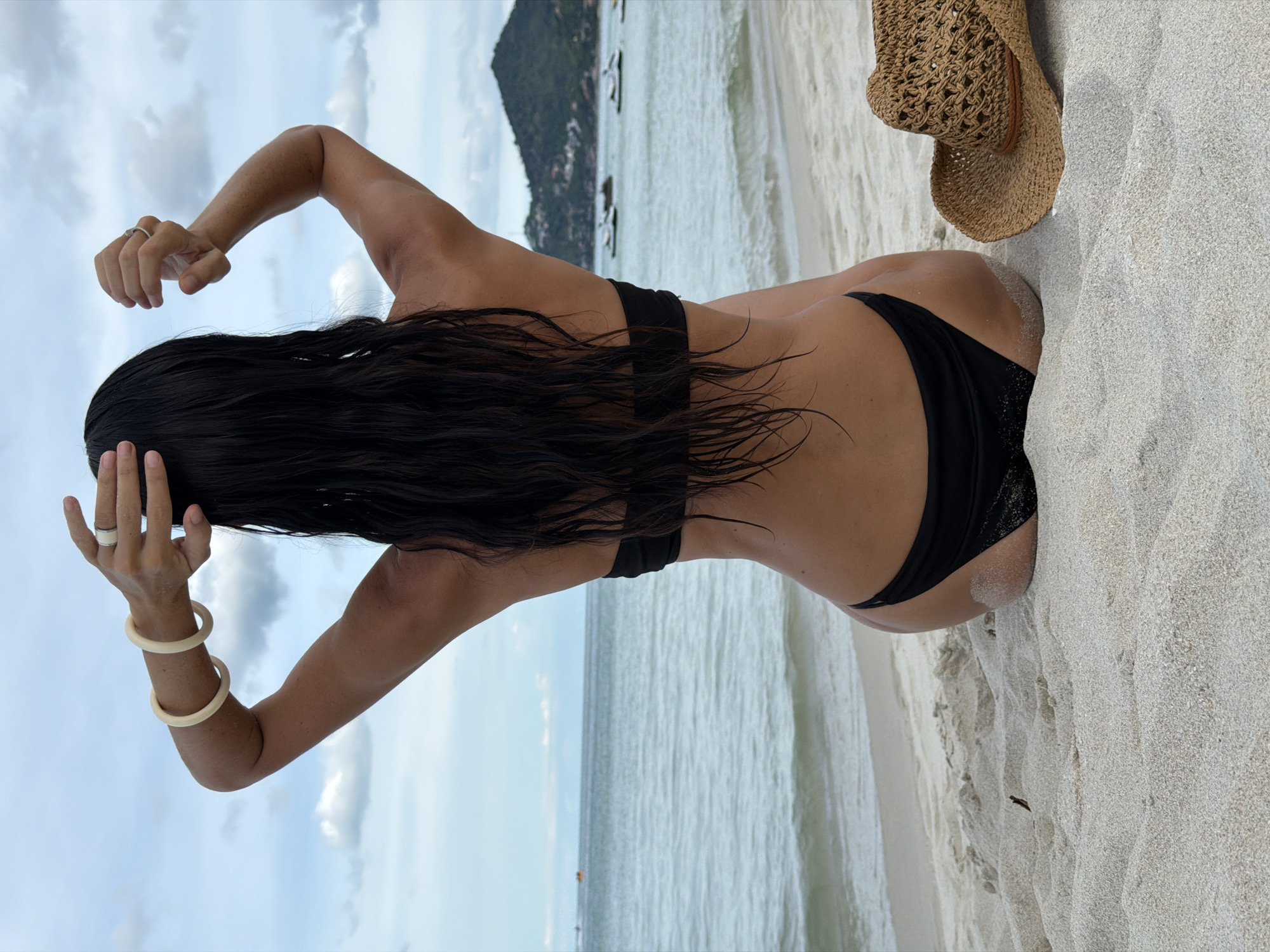 A woman is photographed from behind, sitting on a sandy beach. She has long, dark, wet hair and is wearing a black bikini. Her arms are raised and her hands are resting on her head. She is also wearing jewelry: a ring on her left hand and two bangles on her left wrist. To her right, a straw hat rests on the sand. The background shows the ocean, with a faint outline of a mountainous island in the distance. The sky is cloudy and overcast, suggesting it is daytime, possibly late afternoon or early morning. The overall scene conveys a sense of relaxation and leisure in a tropical beach setting.