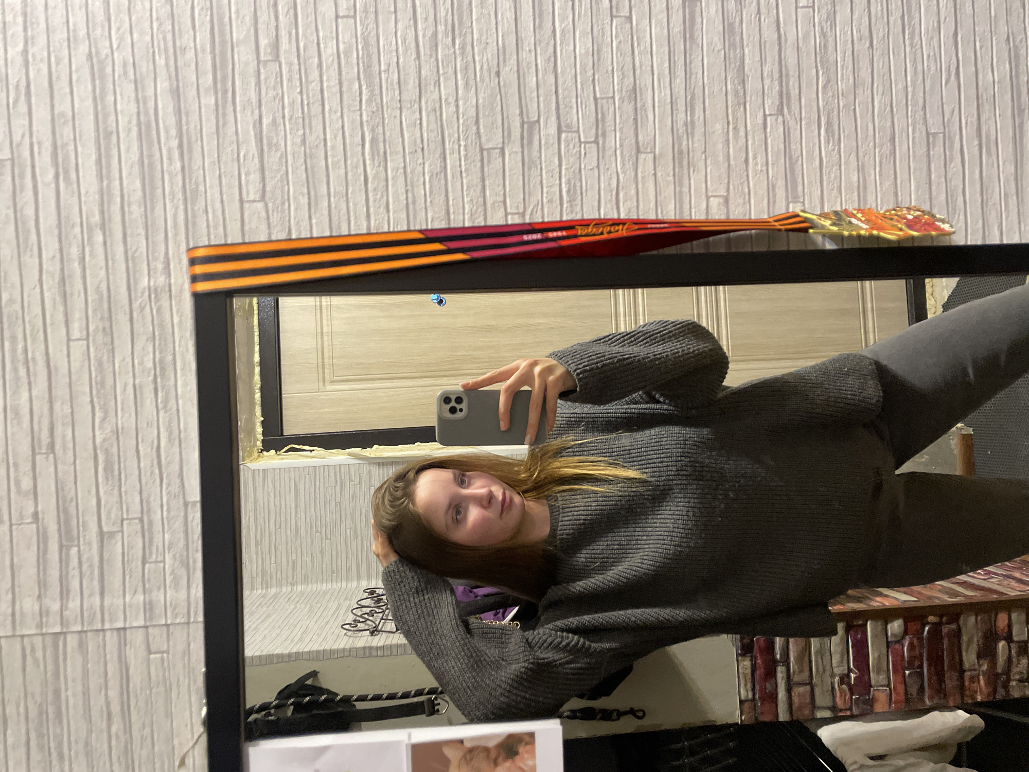 A young adult female is depicted taking a mirror selfie in a residential interior located in Chaikovsky, Russia. She is positioned centrally in the mirror's reflection, looking directly at the smartphone she holds in her right hand, which is visible in the lower right quadrant of the frame. Her left hand is raised, with fingers touching her light brown hair, which is styled with a side part and falls over her shoulders. She is wearing a dark grey, ribbed, long-sleeved sweater and dark grey pants.
The background visible in the mirror's reflection features a light-colored wooden door with paneling details on the right side of the frame. To the left of the door, a section of wall with light grey brick-patterned wallpaper is visible, reflecting behind the subject. Below this, a darker section of wall with red and brown patterned wallpaper or exposed brick is present. A decorative metal scrollwork item and a dark strap or bag are visible behind her on the left. A white piece of paper with an image, possibly of a cat, is taped to the upper left corner of the mirror frame or an adjacent surface.
Outside the mirror's reflection, to the right of the mirror frame, a wall with the same light grey brick-patterned wallpaper extends. Draped over the top right corner of the black mirror frame is a commemorative ribbon featuring prominent black and orange stripes (St. George's ribbon pattern) leading into a red and pink section. White text "Подарок 1945 - 2020" is legible on the red section, signifying a "Gift" related to the 75th anniversary of Victory Day. A gold-colored medal, depicting a star and wreath design, hangs from this ribbon.